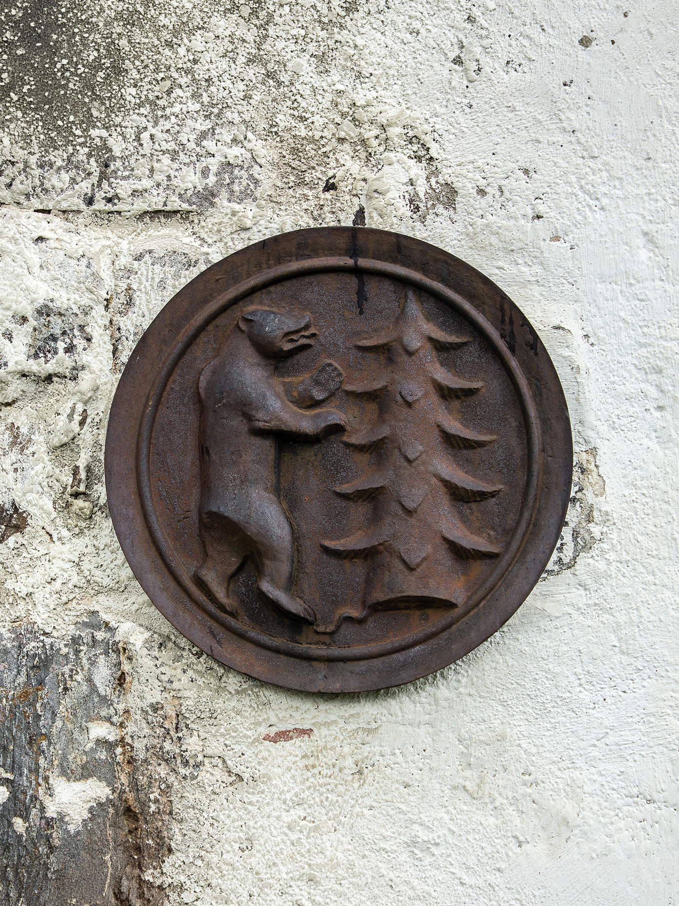Rusty cast iron medallion on a weathered stone wall, depicting a bear and pine tree.