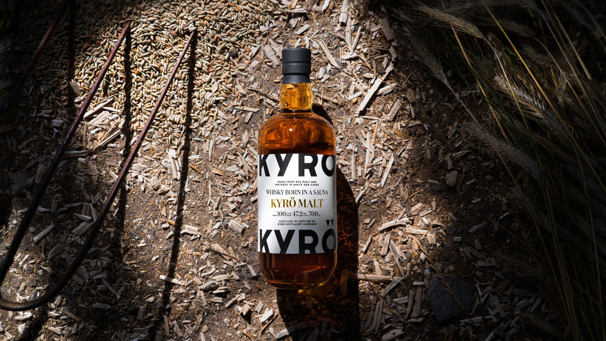 Kyrö Malt whisky bottle resting on wood chips and rye grain, dramatically lit against a dark natural background.