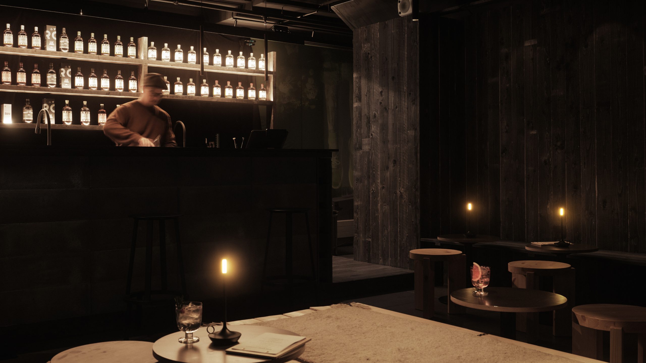 Kyrö Distillery bar interior at night, backlit whisky bottles on shelves, candlelit tables, dark wood walls, bartender in motion.