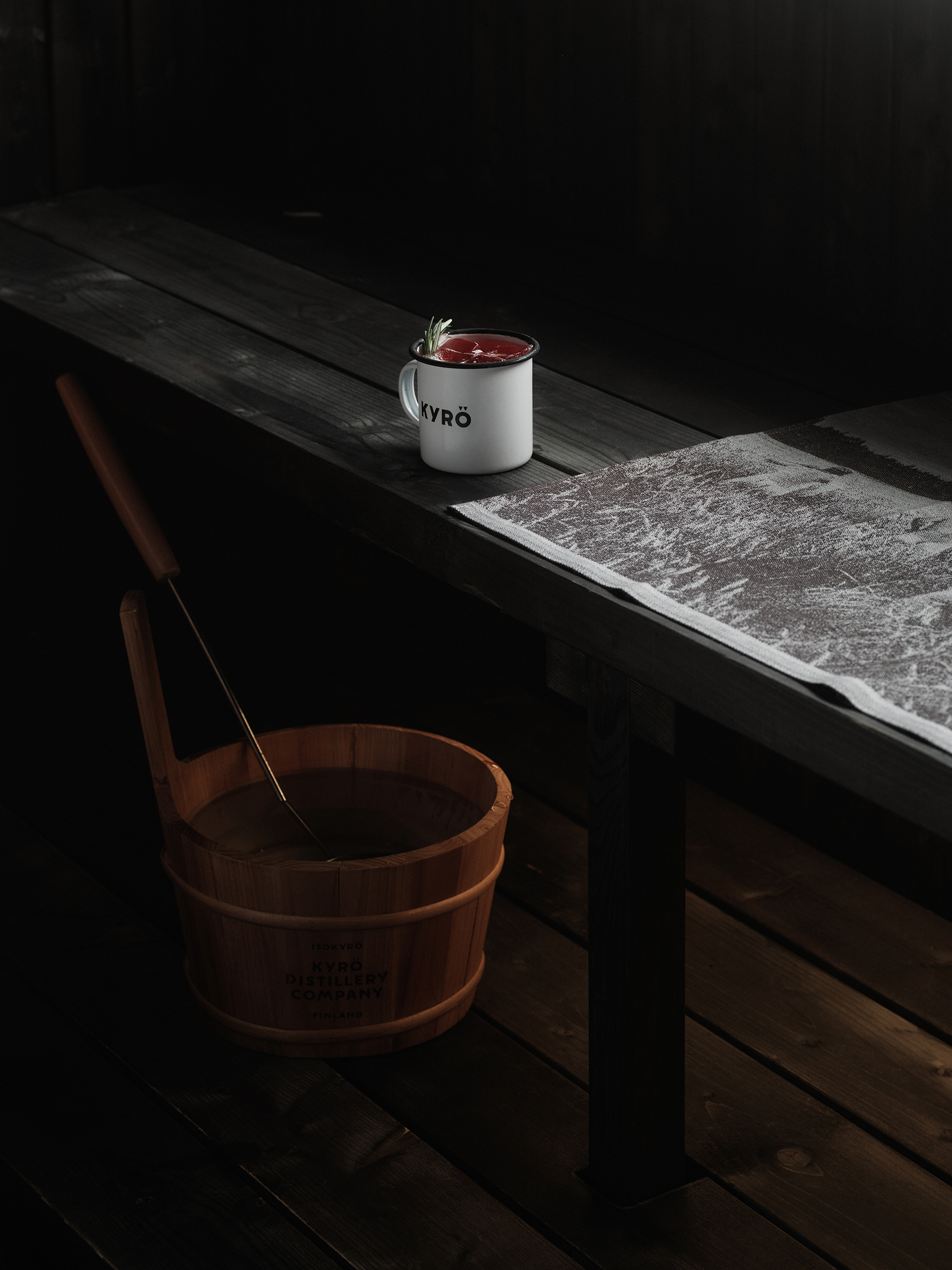 Kyrö enamel mug with red cocktail on dark sauna bench beside a branded wooden bucket and ladle.