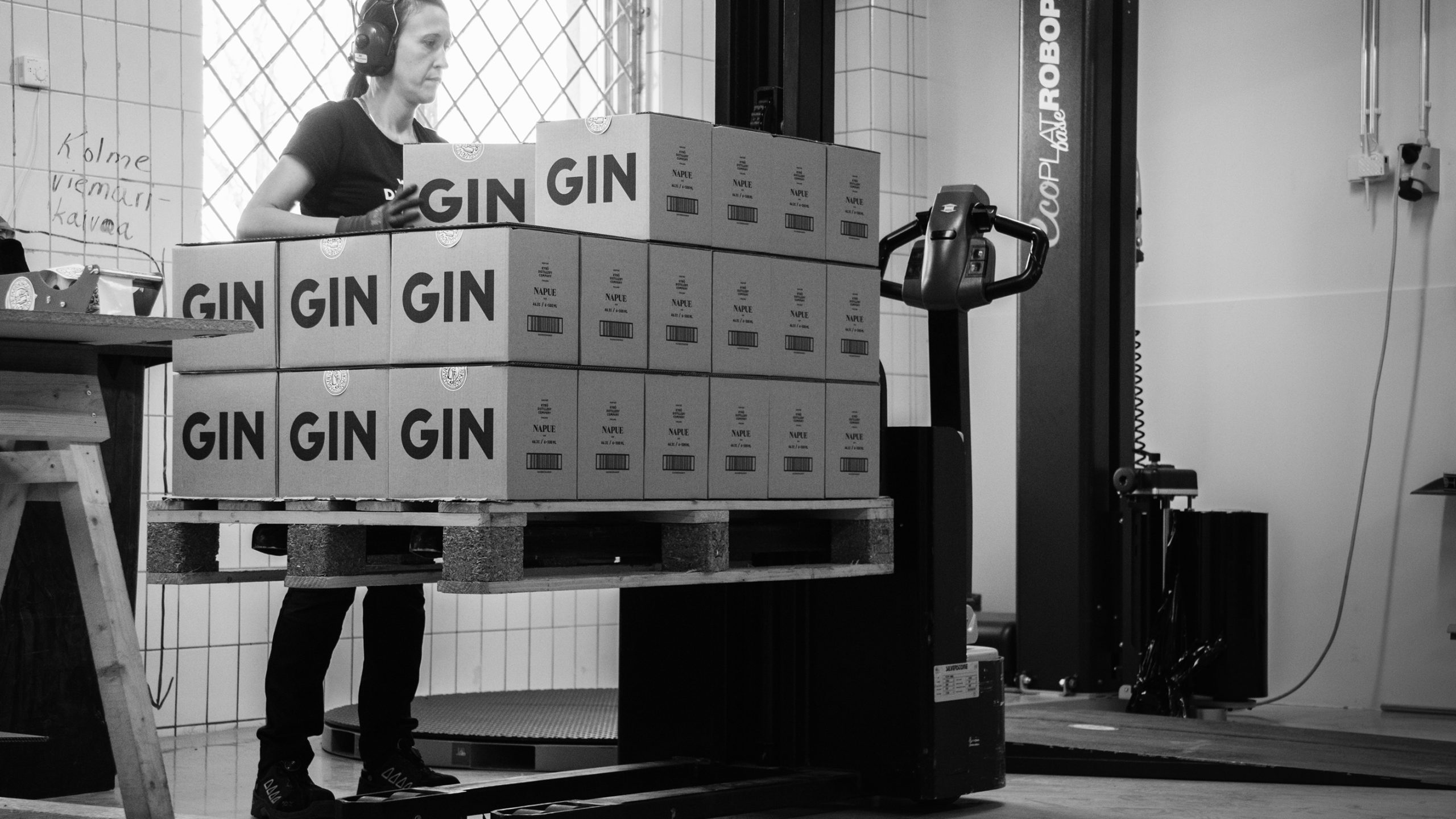 Worker stacking Kyrö Distillery GIN boxes on a pallet in a warehouse, wearing headphones, next to an automated wrapping machine.