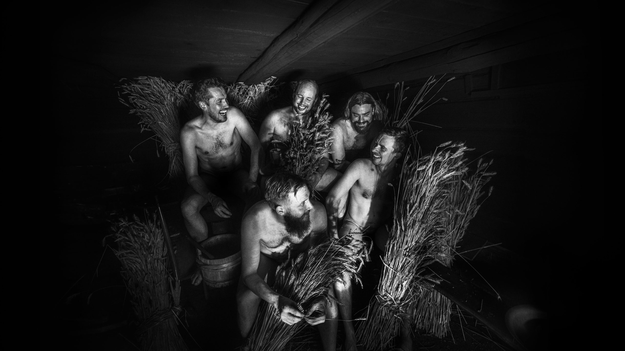 Five laughing men in a smoke sauna surrounded by grain stalks and a wooden bucket, evoking Kyrö Distillery's Finnish heritage.