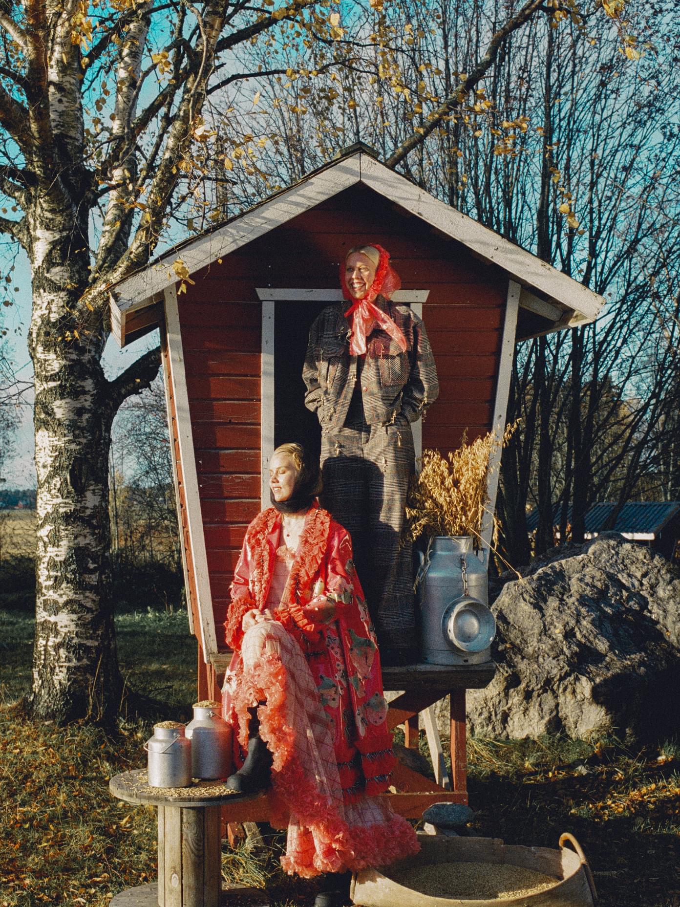 Two models in bold red and plaid outfits pose at a rustic red wooden shed with milk churns for Mö branding by Werklig.
