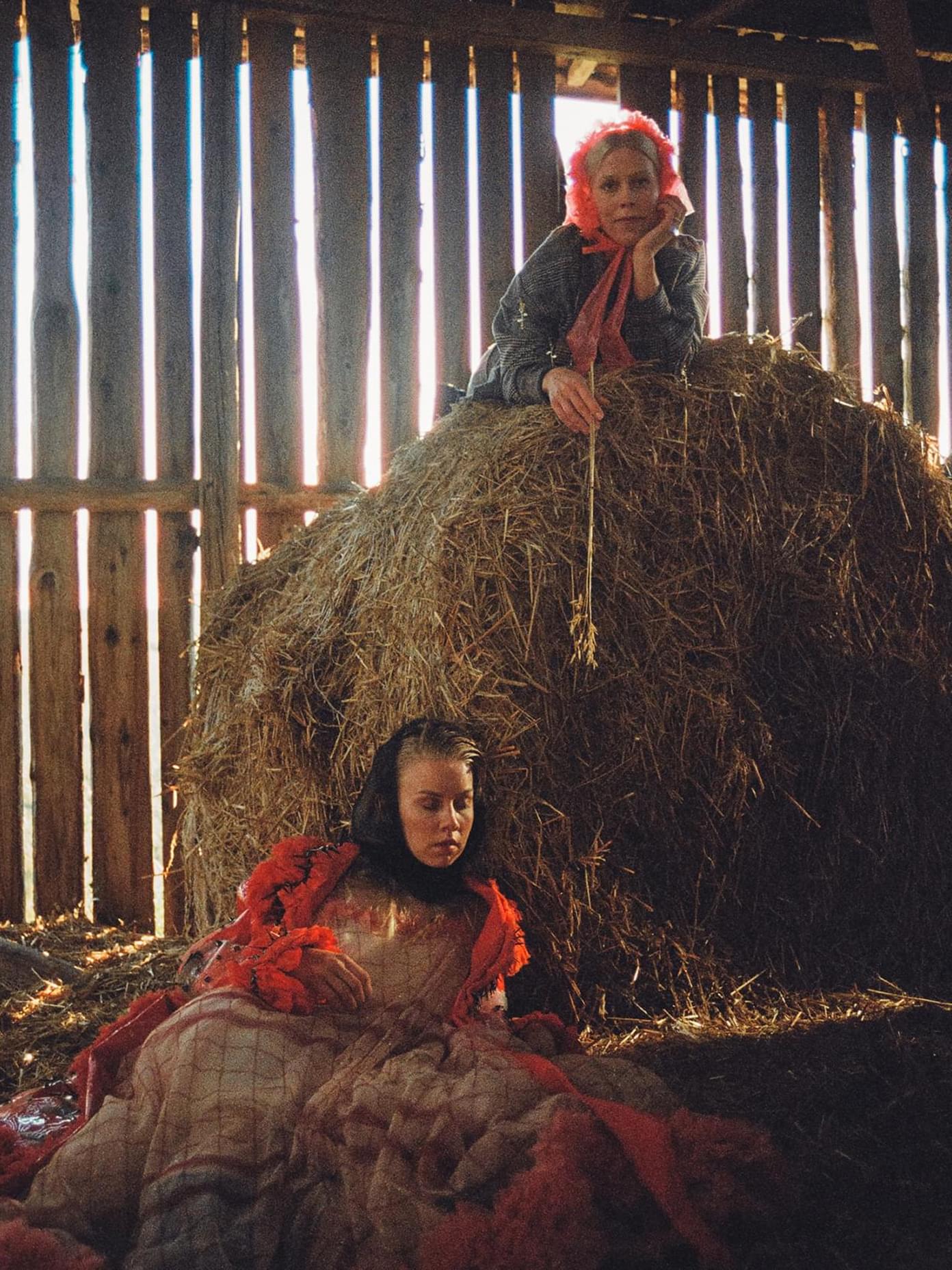 Two women in a rustic barn with a hay bale, wearing red and grey garments, in a moody editorial scene for Mö.