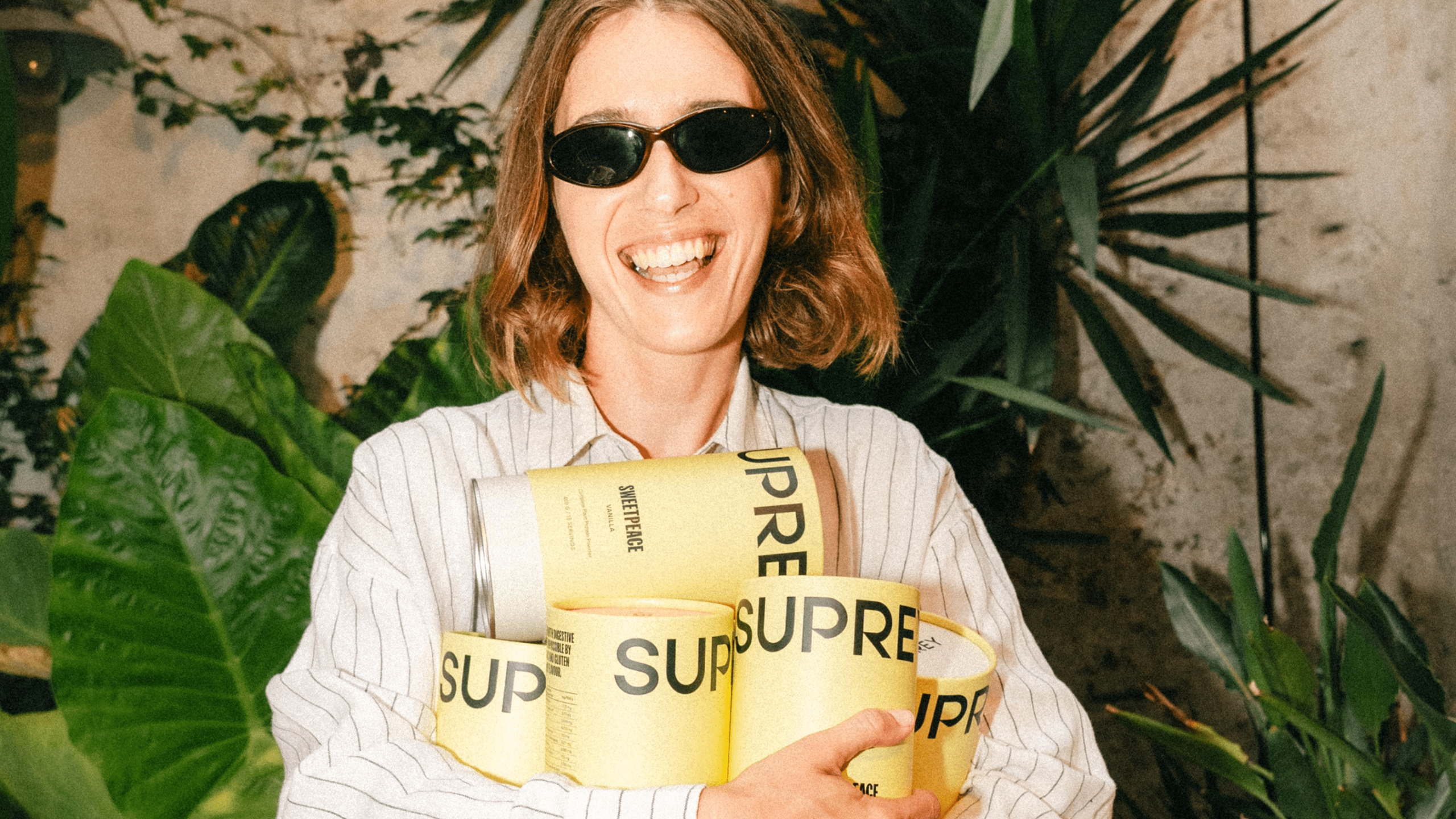 Smiling person holding multiple yellow Suprey supplement canisters, wearing sunglasses and a striped shirt, surrounded by tropical plants.