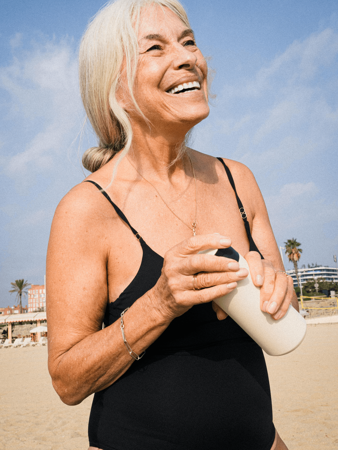 Smiling older woman in black swimsuit holding Suprey sunscreen on a sunny beach, radiating confidence and joy.