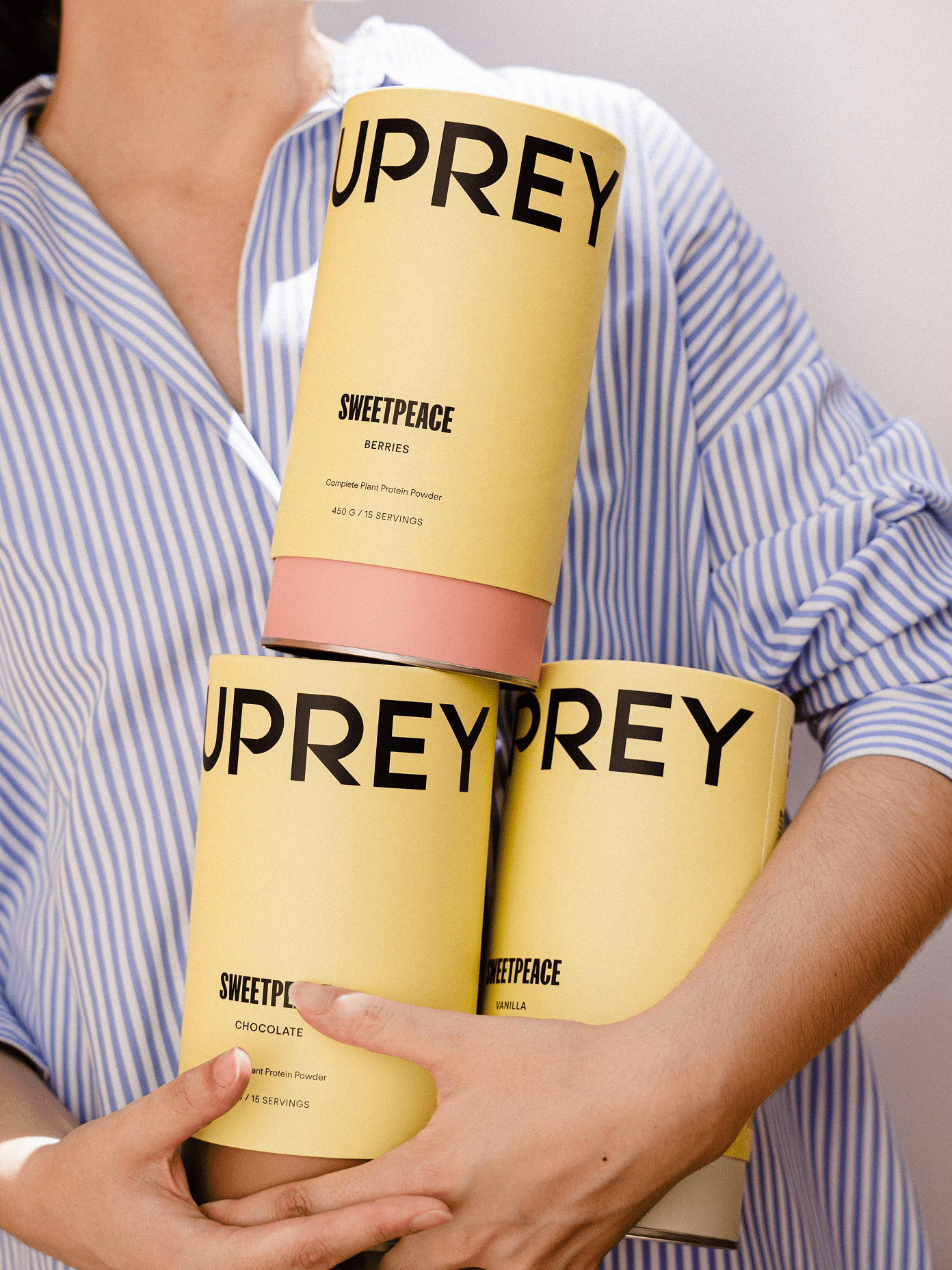 Person in blue striped shirt holding three yellow cylindrical Suprey Sweetpeace plant protein powder containers in berries, chocolate, and vanilla.