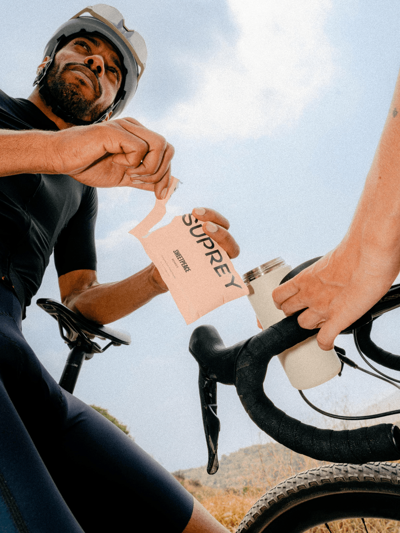 Cyclist opening a Suprey nutrition packet while another rider holds handlebars with a water bottle, shot from below.