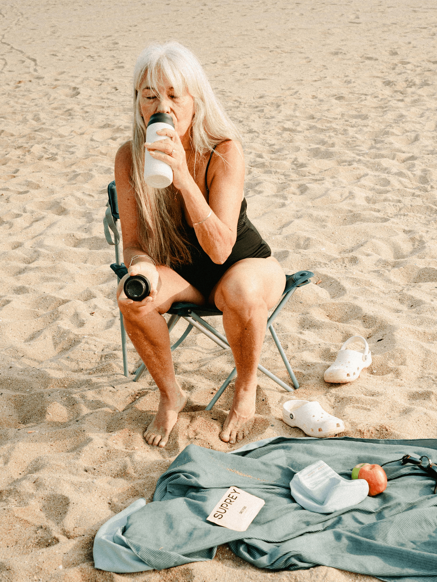Older woman with long white hair drinking from a Suprey bottle on a beach chair, Crocs and a teal blanket nearby.