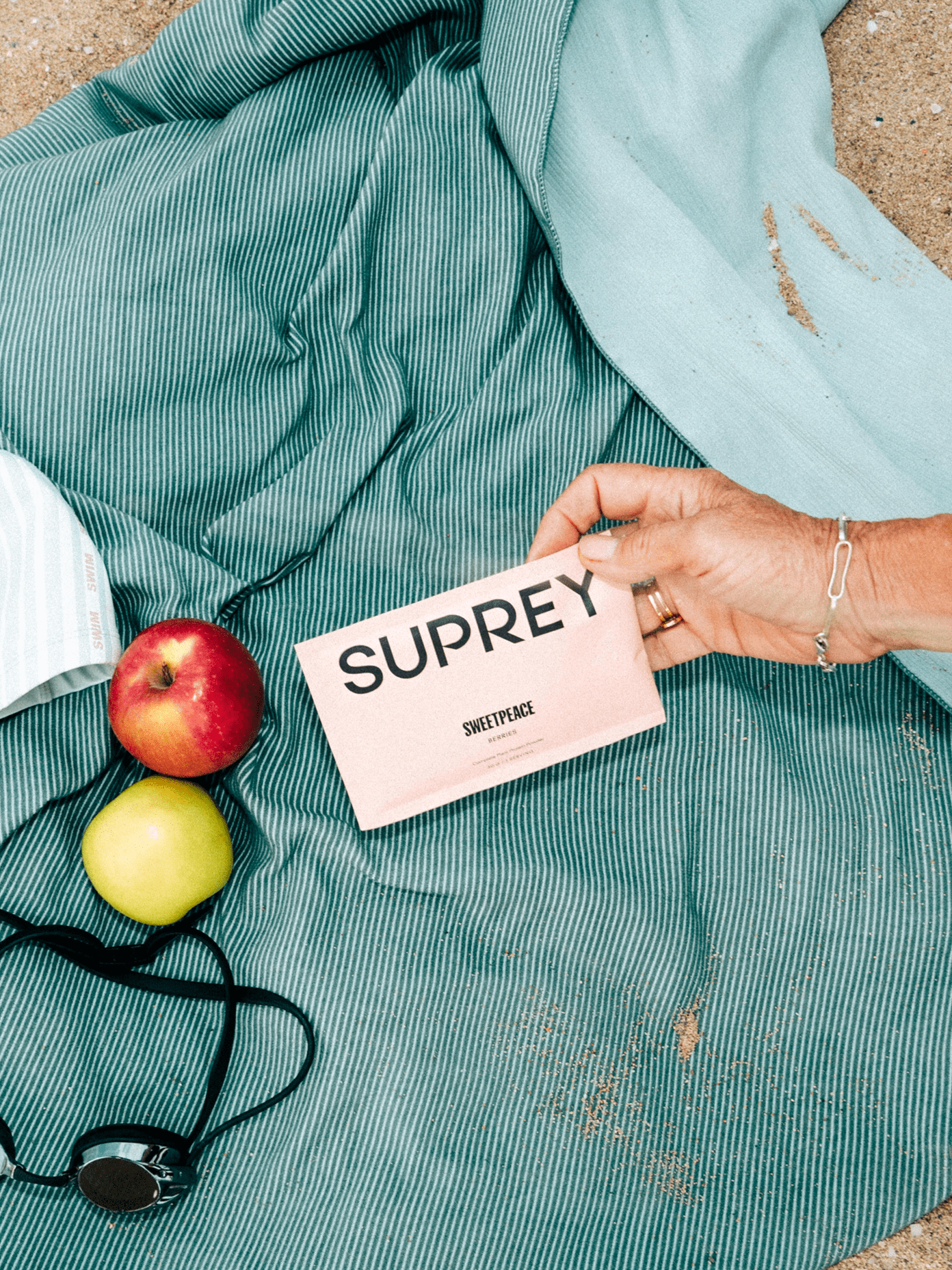 Hand holding a pink Suprey packet on a striped beach towel beside two apples and swim goggles.