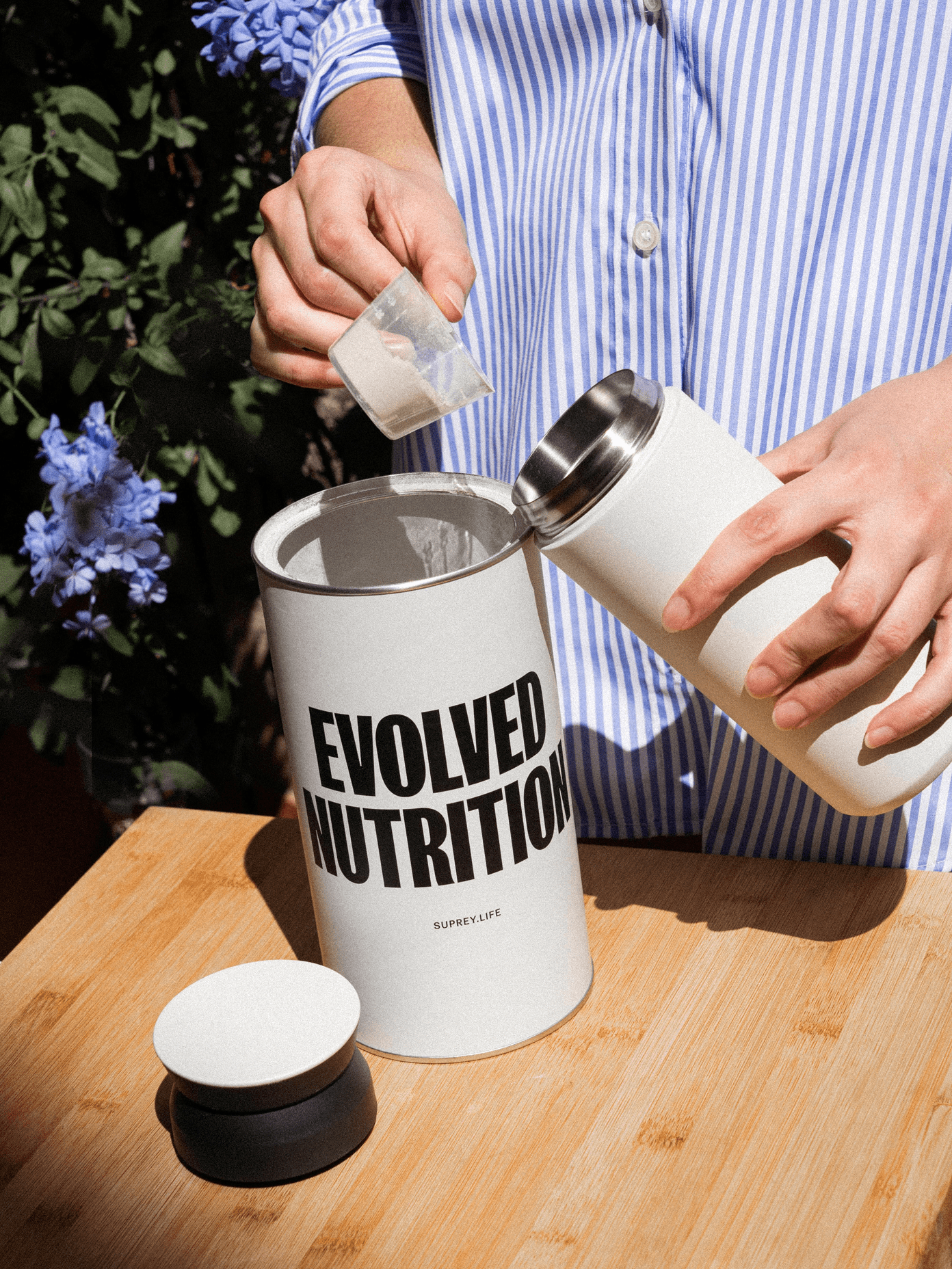 Person in striped shirt scooping powder from an Evolved Nutrition canister by Suprey into a white tumbler outdoors.