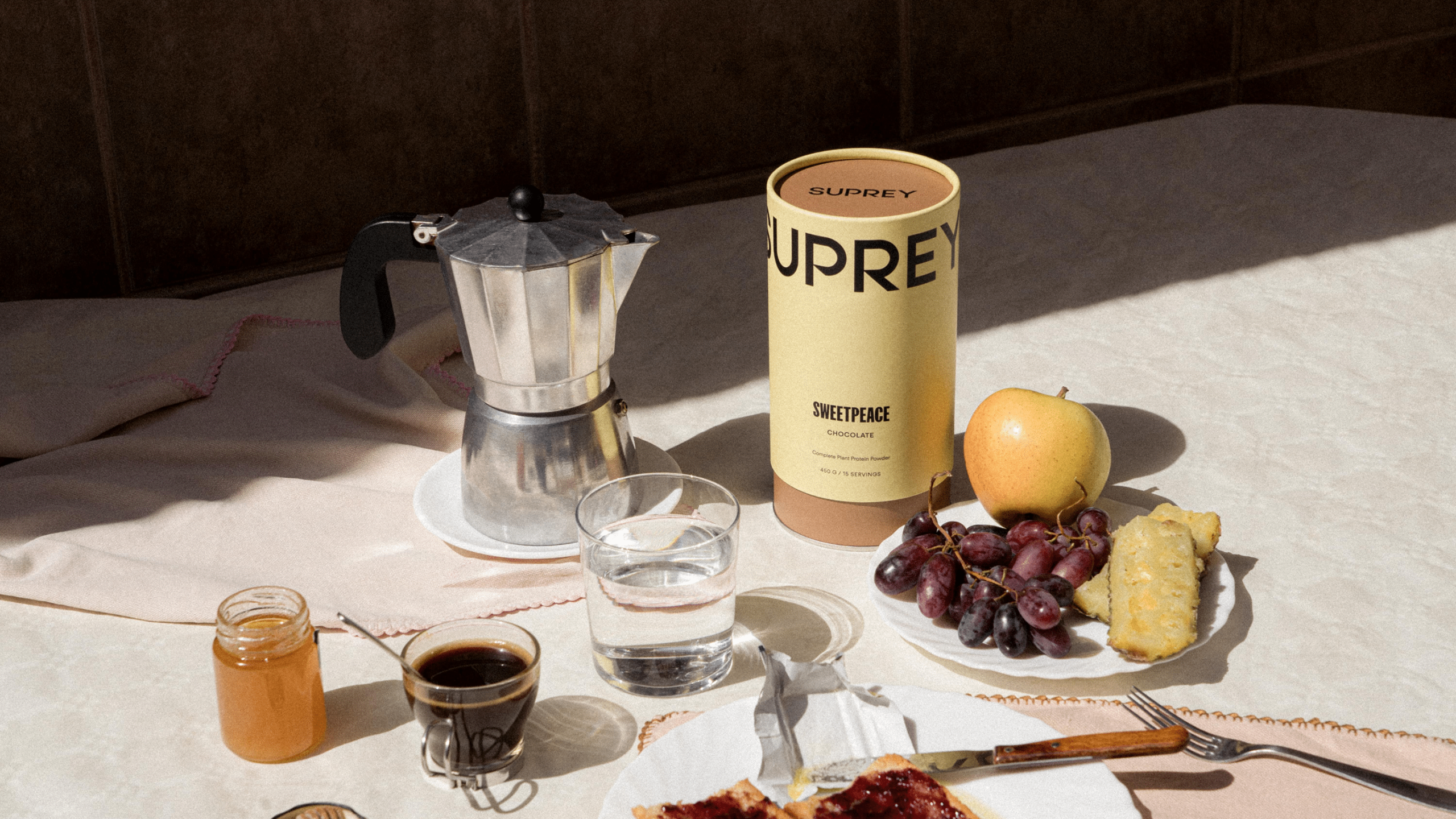 Suprey Sweetpeace chocolate protein powder canister styled on a breakfast table with moka pot, espresso, fruit, and toast.
