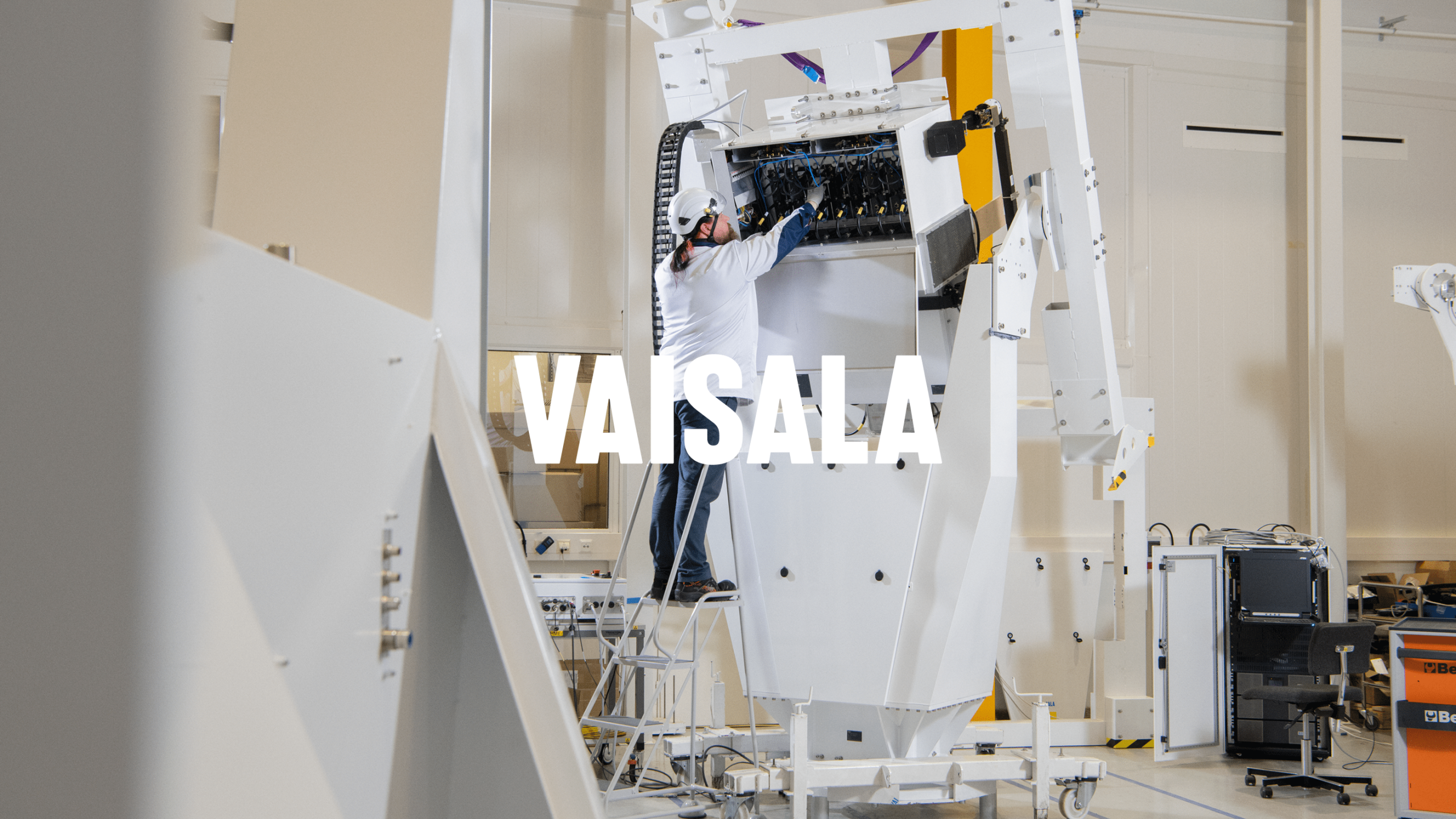 Vaisala technician in white hardhat works on large white industrial equipment inside a high-tech facility.