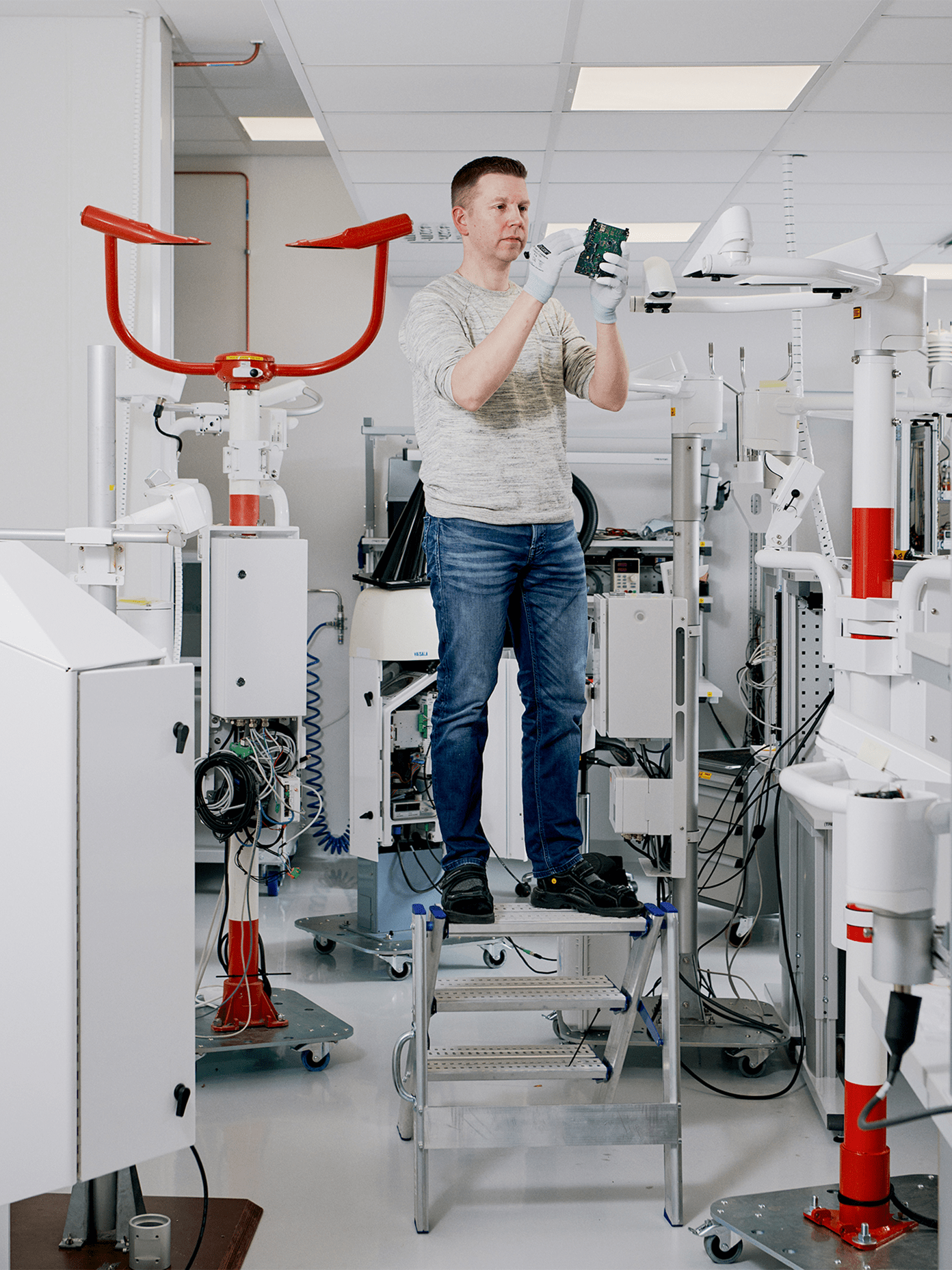 Vaisala technician in white gloves stands on a step ladder inspecting a circuit board amid white laboratory equipment.