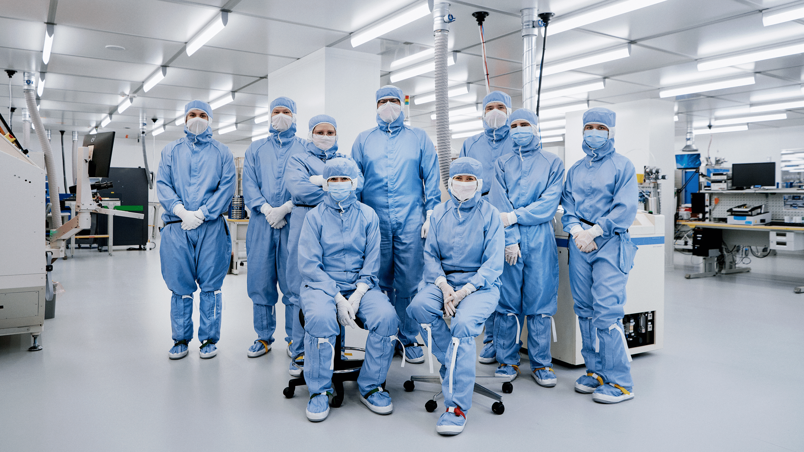 Vaisala cleanroom team of nine wearing blue protective suits, masks, and gloves posing in a bright semiconductor facility.