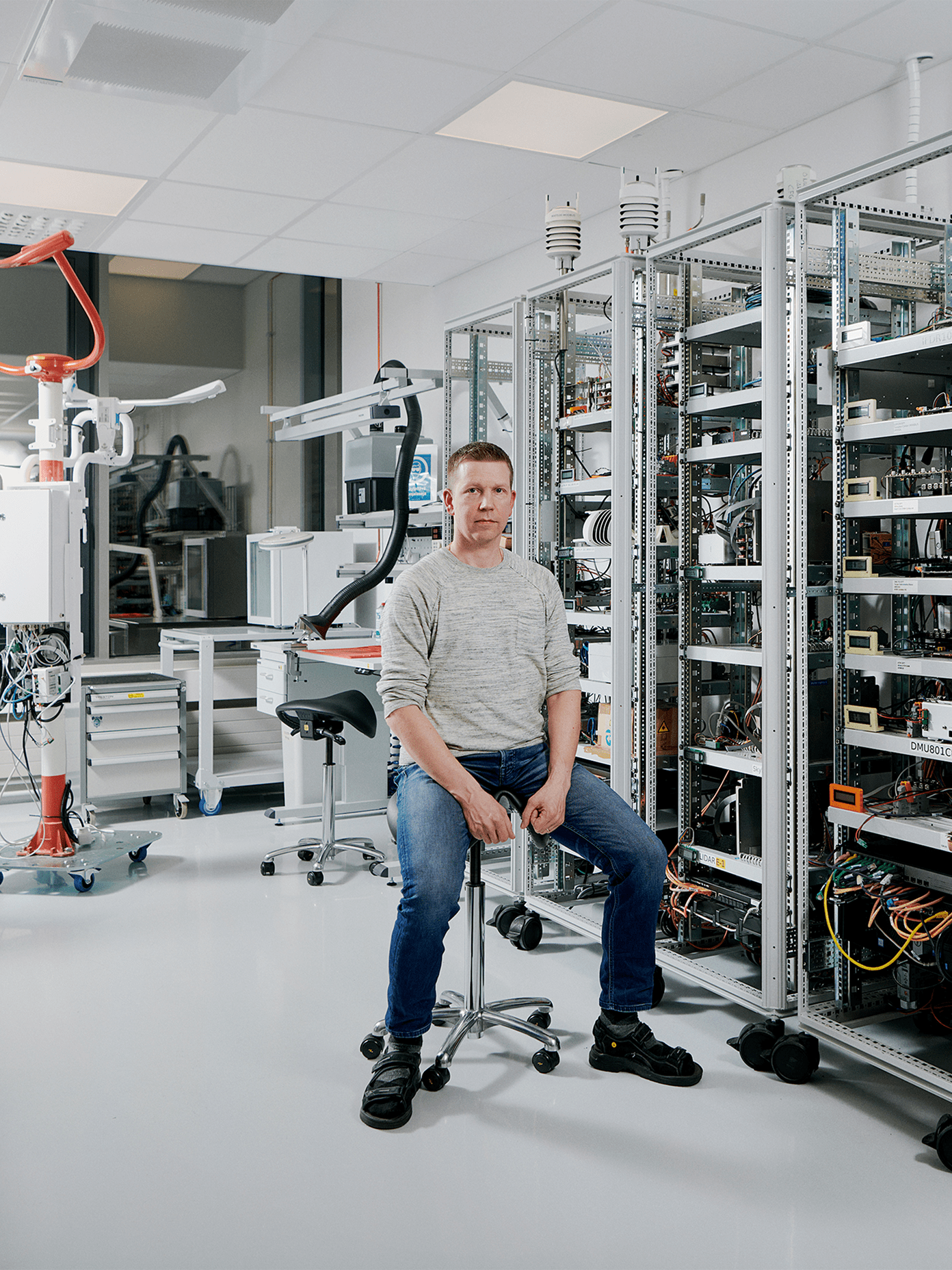 Vaisala engineer seated on a stool amid tall equipment racks filled with electronics and sensors in a modern laboratory.