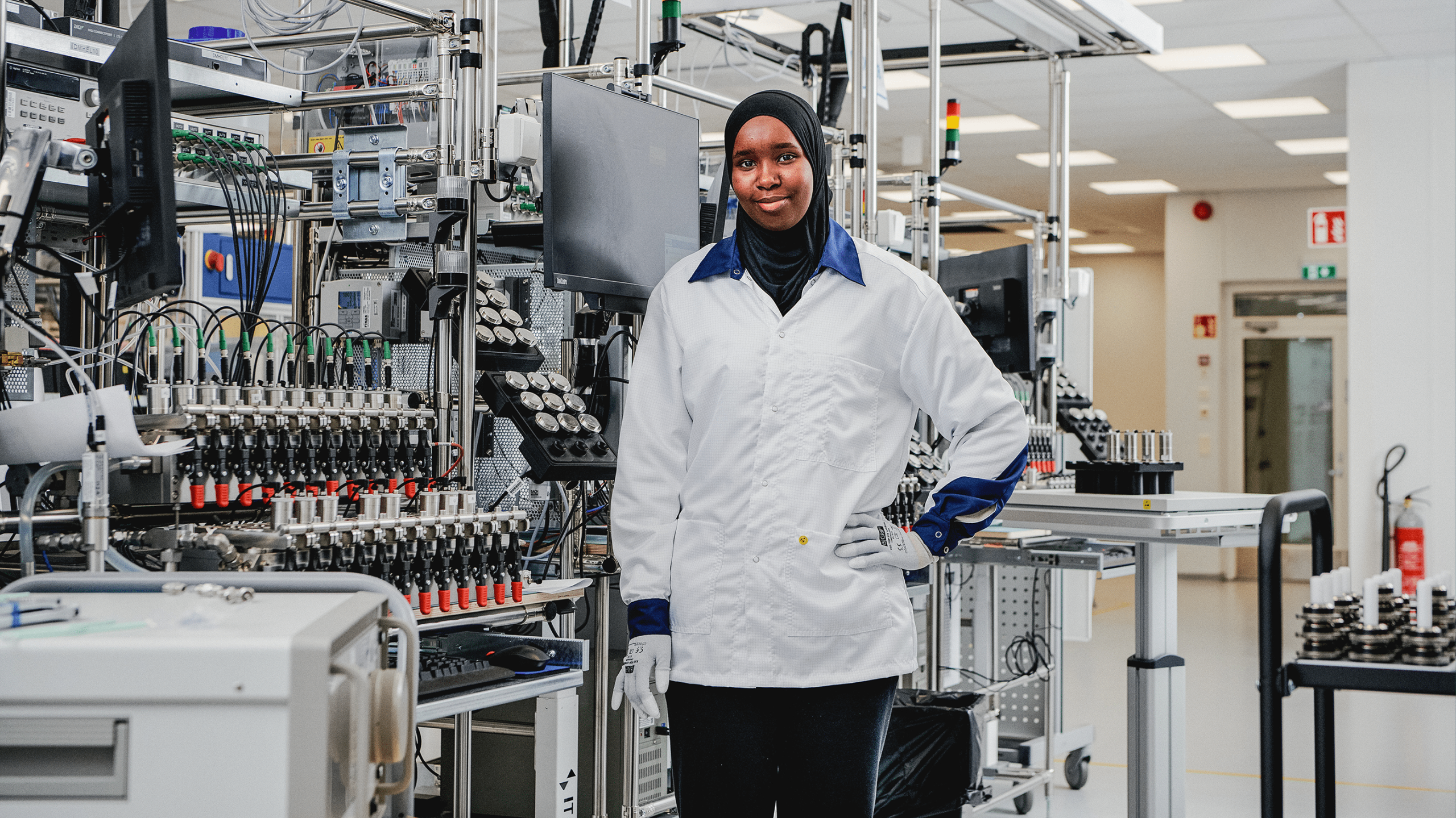 Vaisala technician in white lab coat and hijab stands confidently among complex electronic testing equipment on a factory floor.