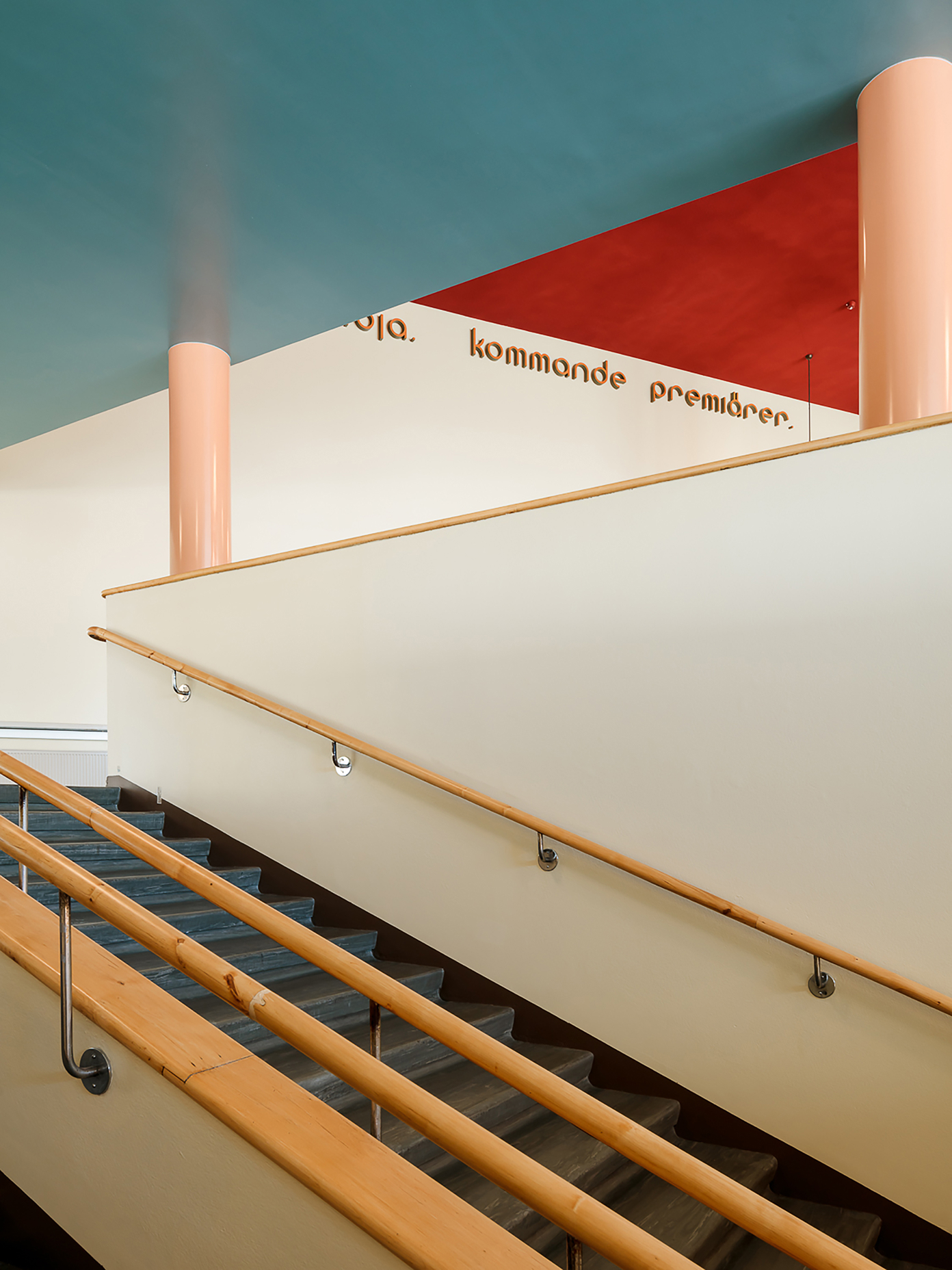 Interior staircase at Amos Rex with a teal ceiling, red accent wall, and cream handrail, Finnish exhibition text visible above.