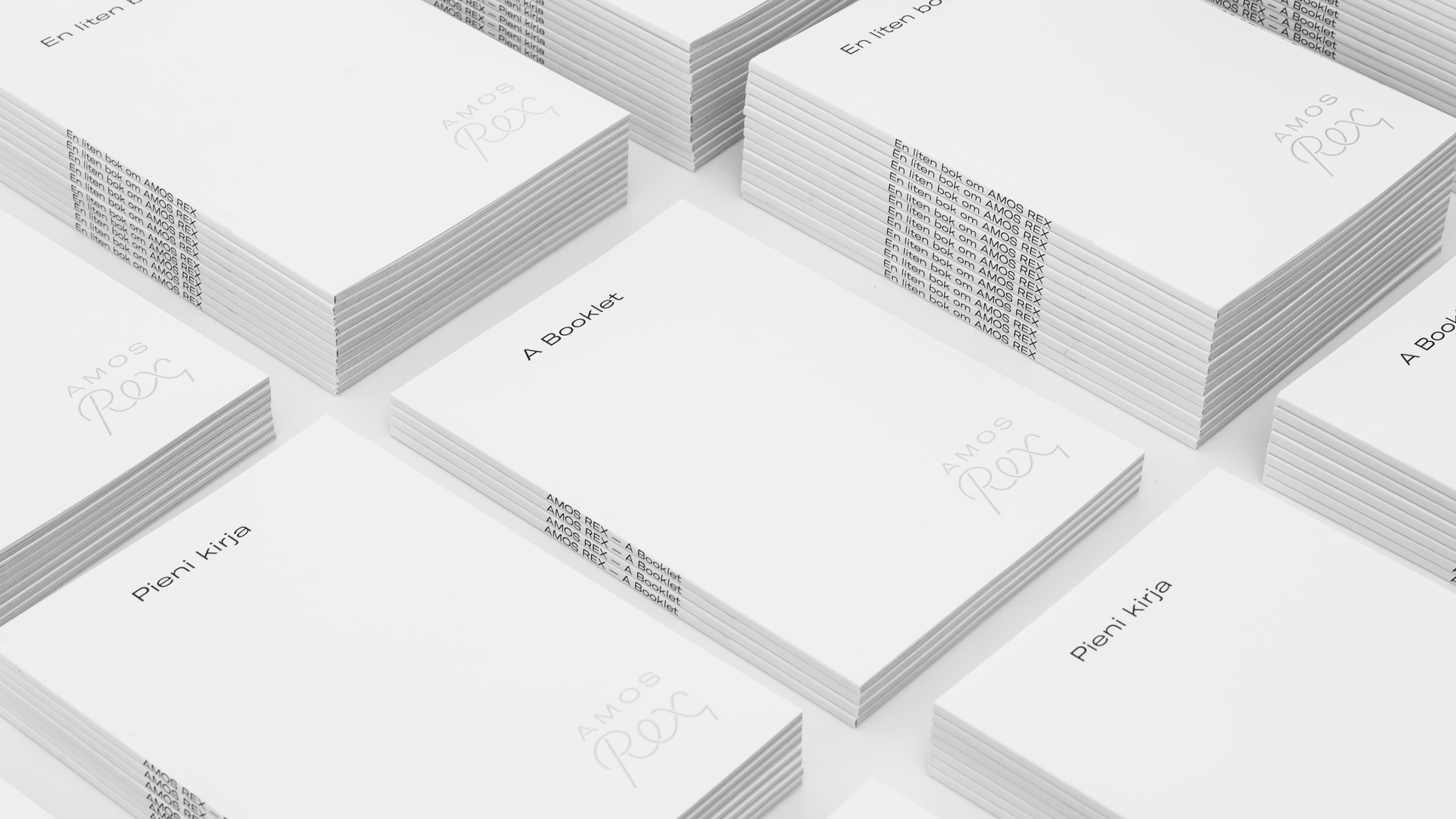 Stacks of white Amos Rex booklets in Finnish, Swedish, and English, each embossed with the museum's script wordmark.