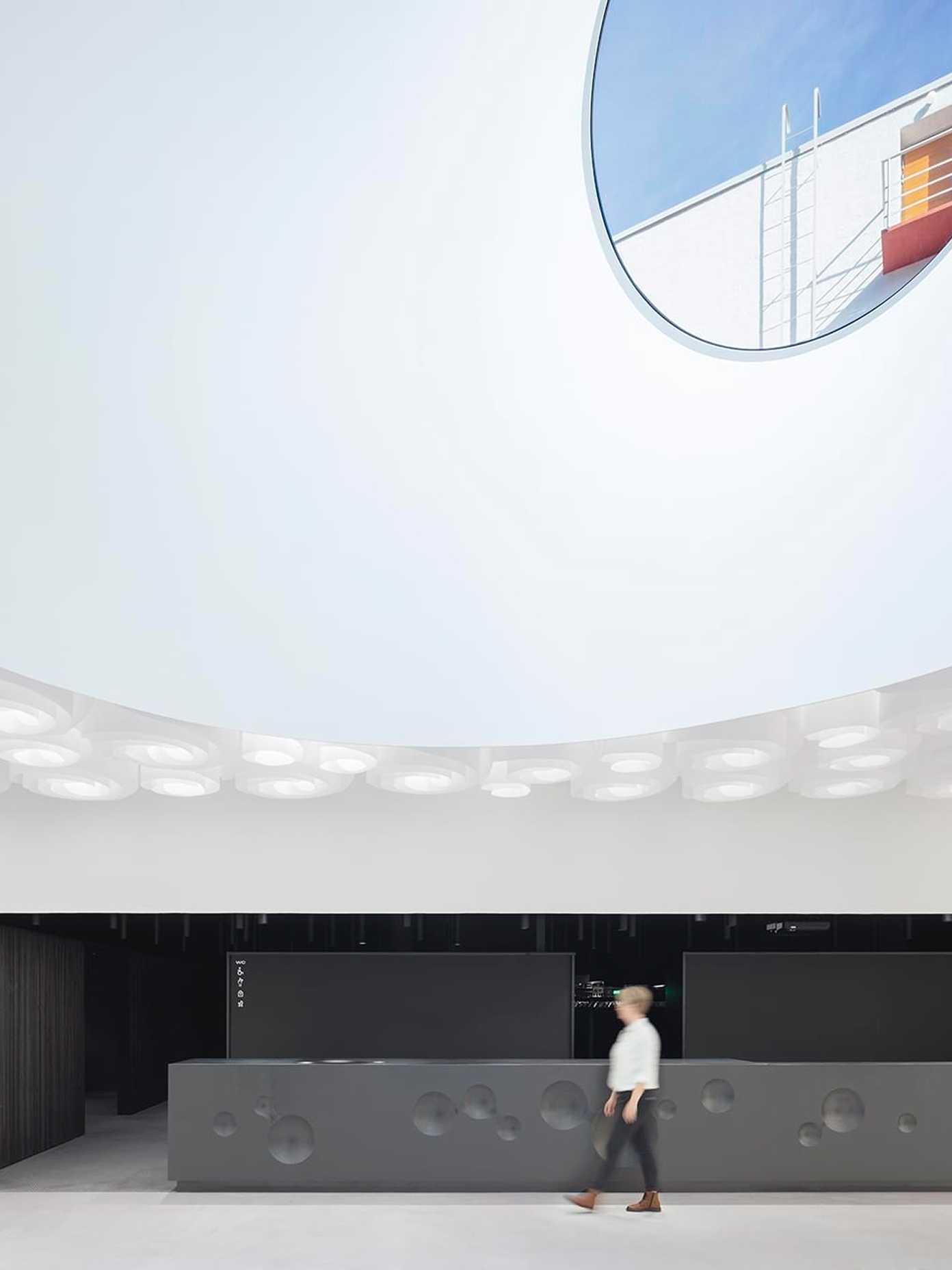 View up through Amos Rex's circular skylight to the sky, above the museum's dark perforated reception desk where a visitor walks past.