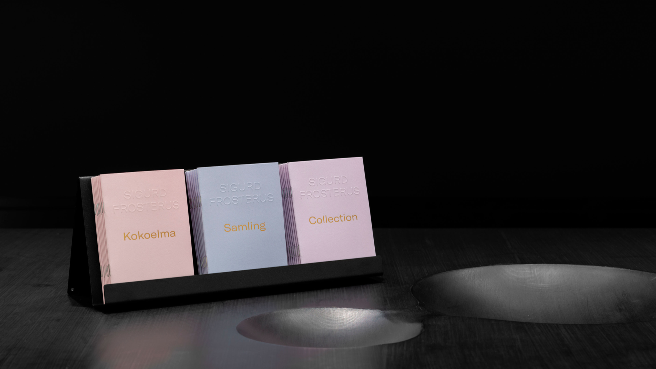 Three Sigurd Frosterus collection booklets in pink, lavender, and light pink, titled in Finnish, Swedish, and English, displayed in a stand.