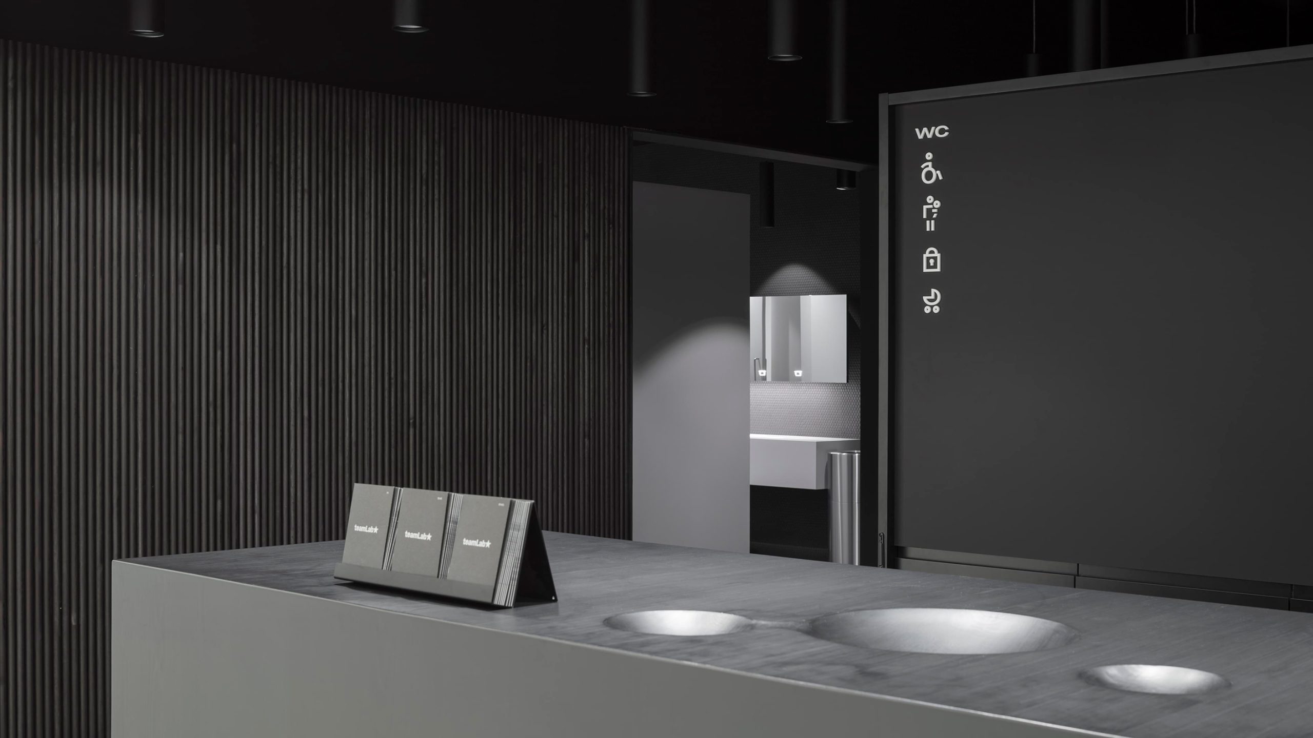 Amos Rex restroom signage with accessibility icons beside a reception desk displaying dark teamLab exhibition guide booklets.