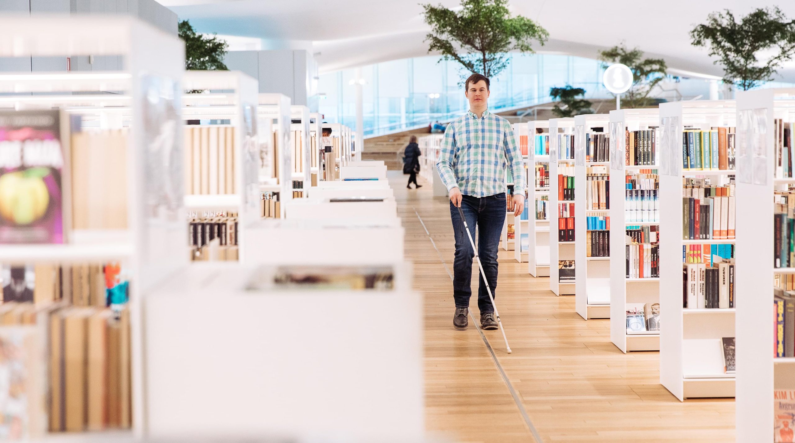 A man using a white cane walks along a tactile guide line between bookshelves at Oodi library.