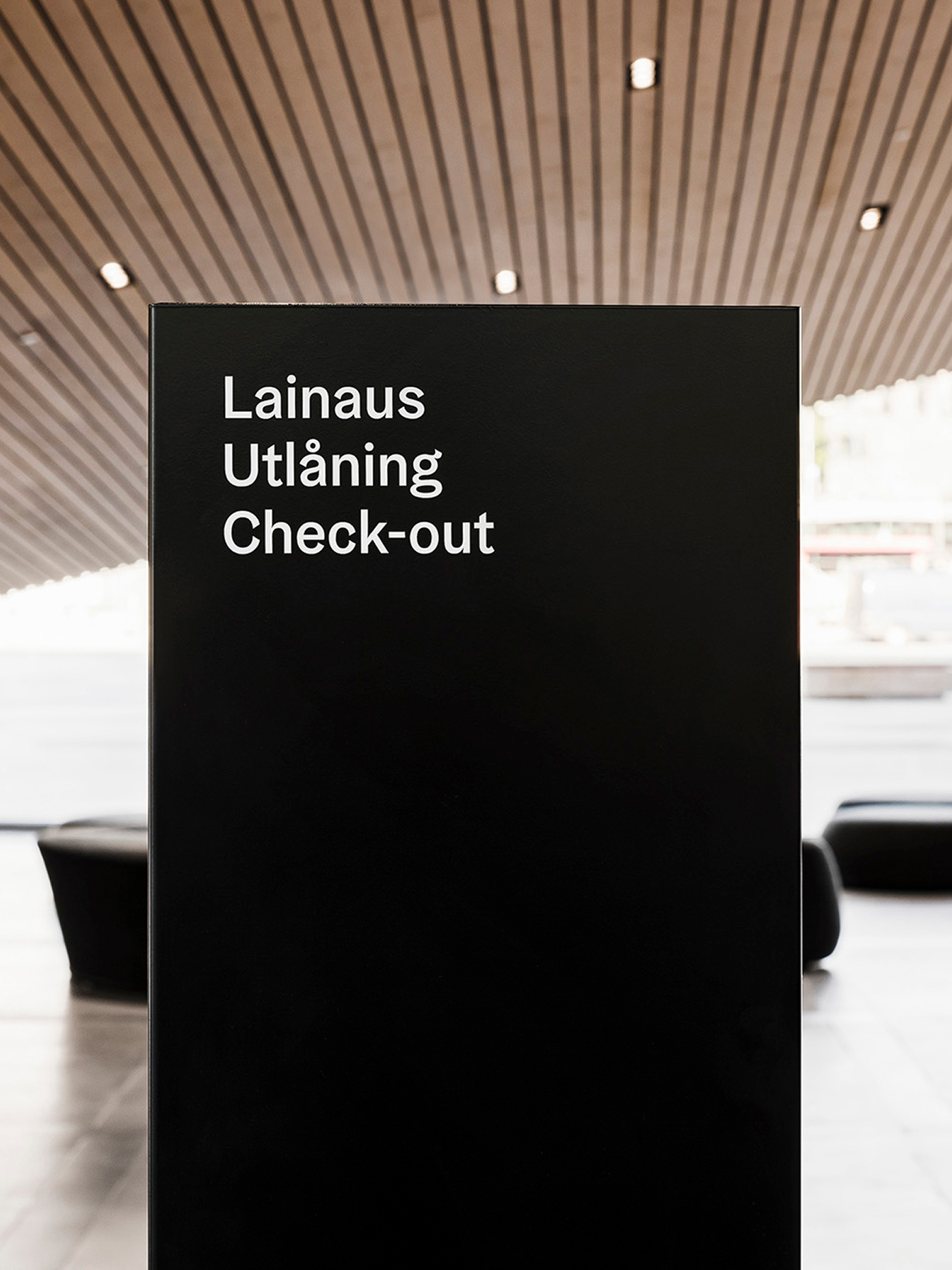 Trilingual 'Check-out' signage in Finnish, Swedish, and English beneath Oodi library's wood-slat ceiling.