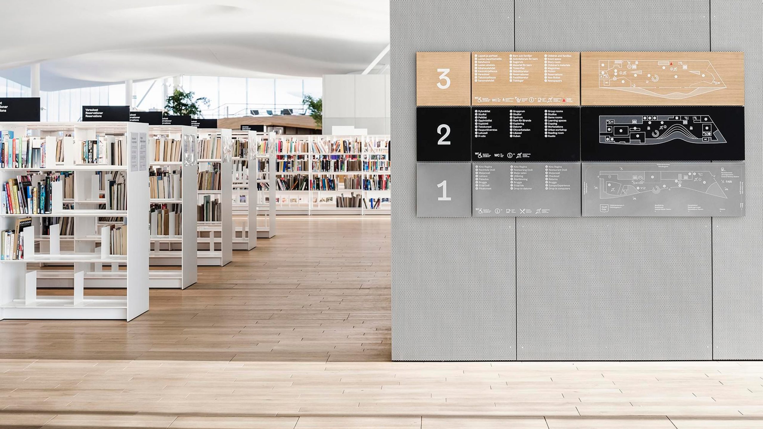 Floor directory signage listing services on levels 3, 2, and 1 with icons and maps, beside Oodi library bookshelves.