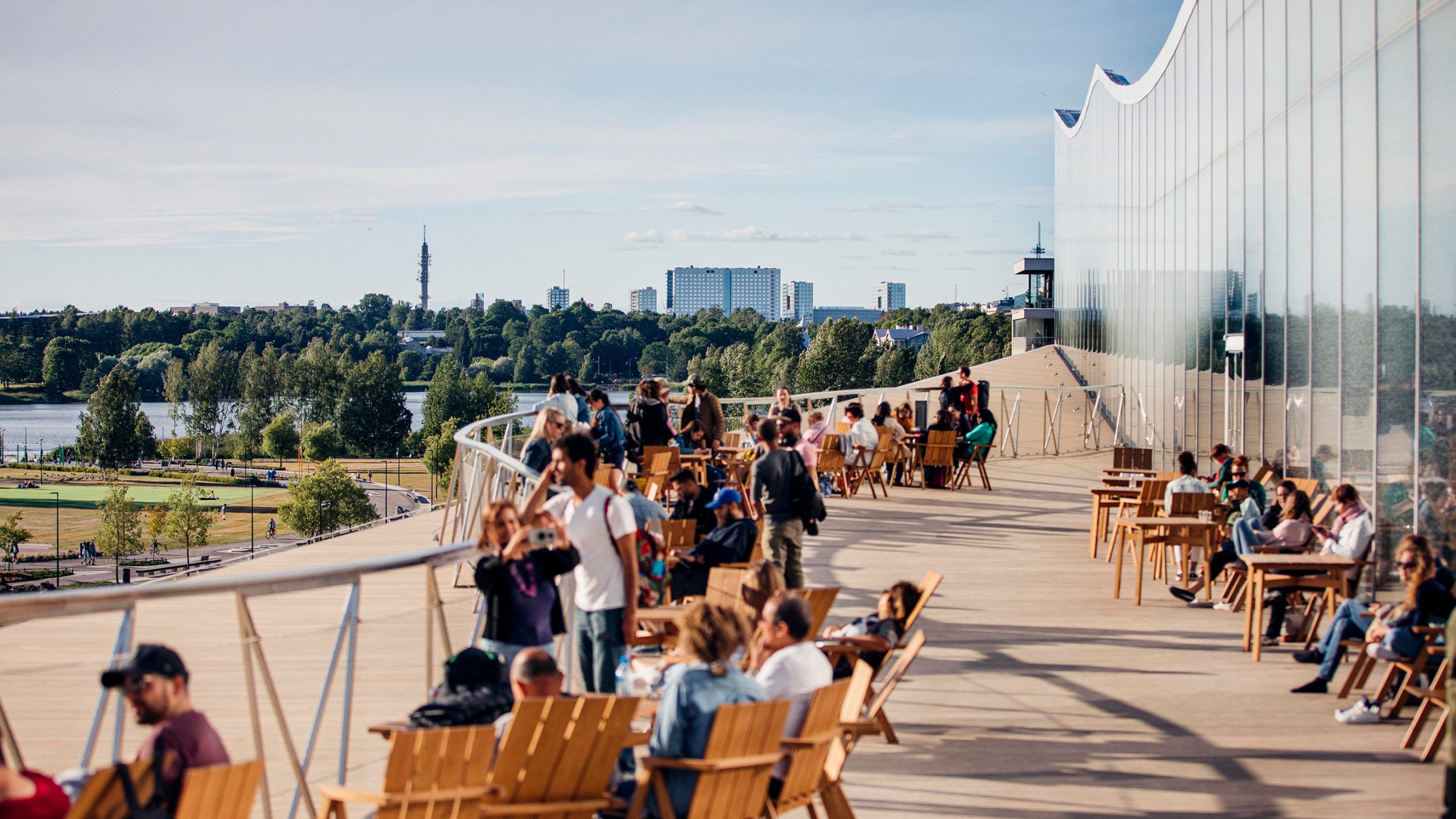 People relaxing at tables on Oodi library's rooftop terrace, with Helsinki's skyline and parkland visible beyond the wavy glass facade.