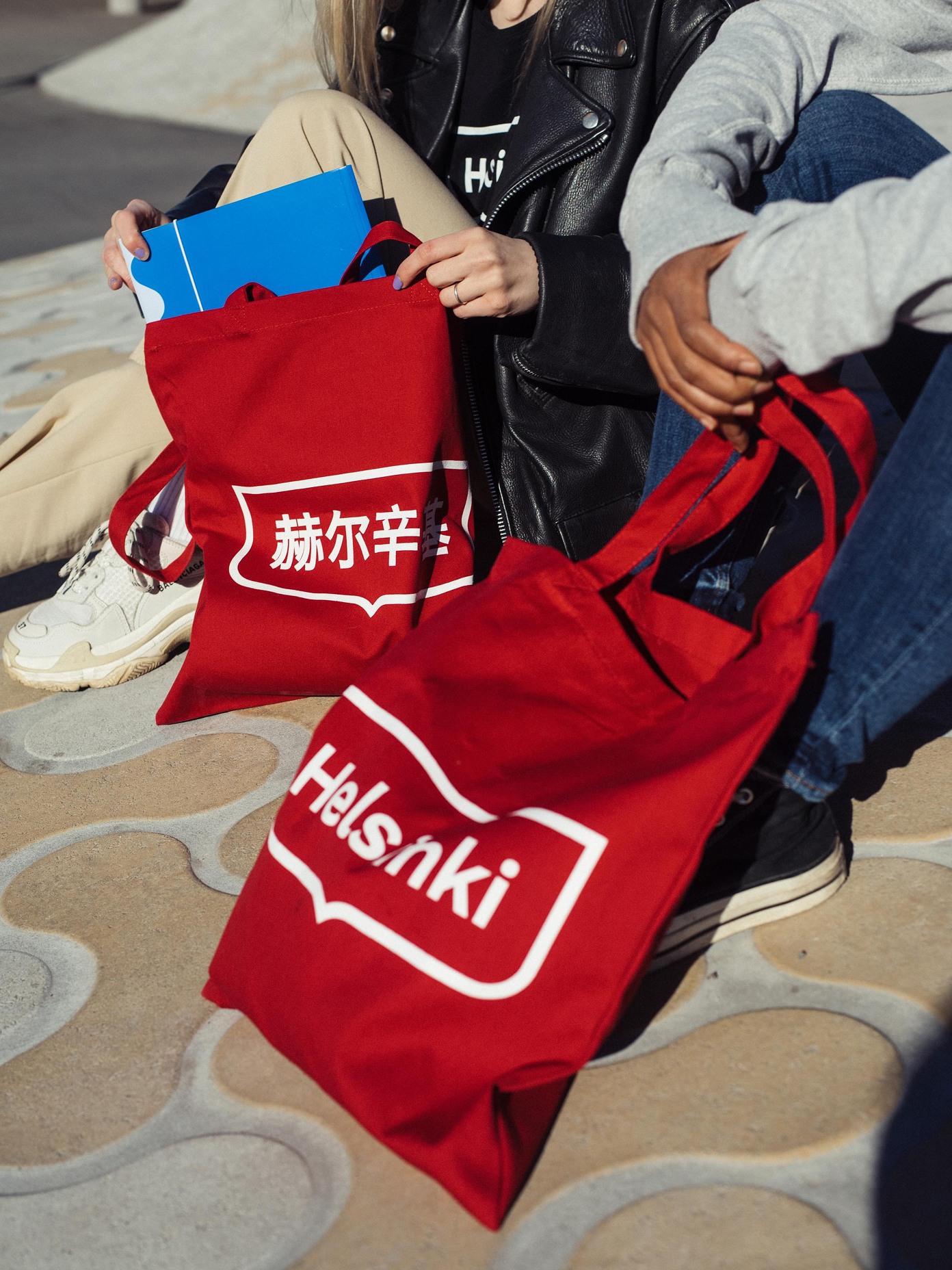 Two people holding red tote bags branded with the Helsinki logo, one in Chinese characters and one in English.