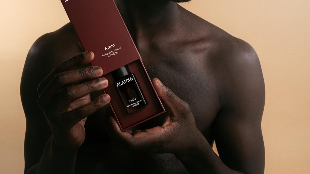Hands holding open a deep red box containing Blank& Amio Refreshing Face Oil with CBD in a dark glass bottle.