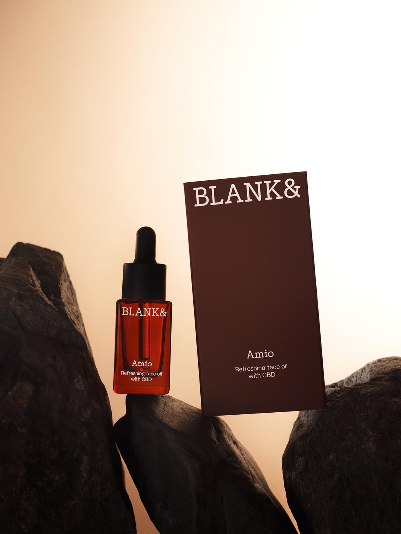 Blank& Amio CBD face oil dropper bottle and dark red packaging box arranged among dark rocks with warm glowing background.