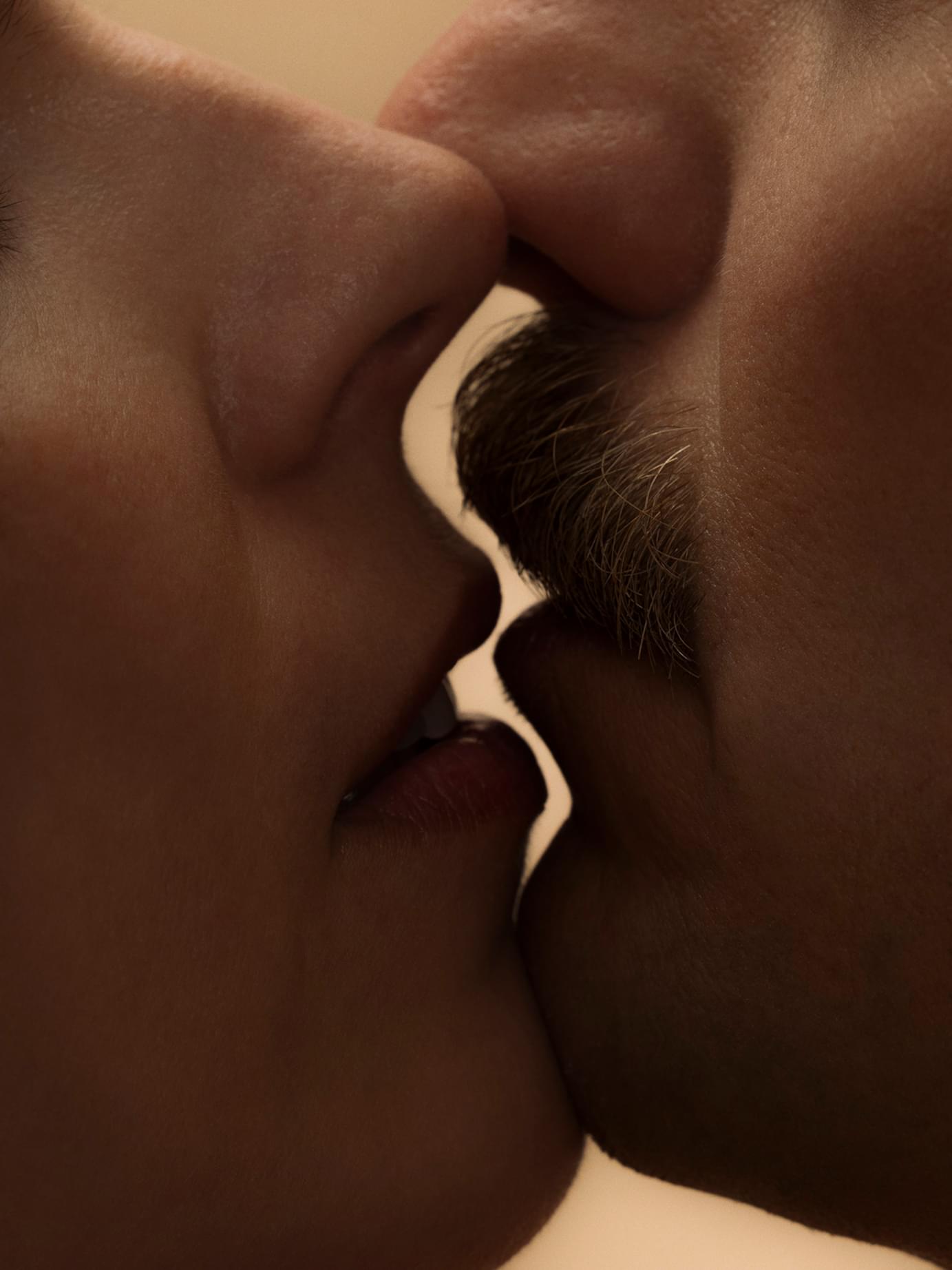 Close-up of two faces almost kissing, with a wavy white line separating them, highlighting facial hair and lips.
