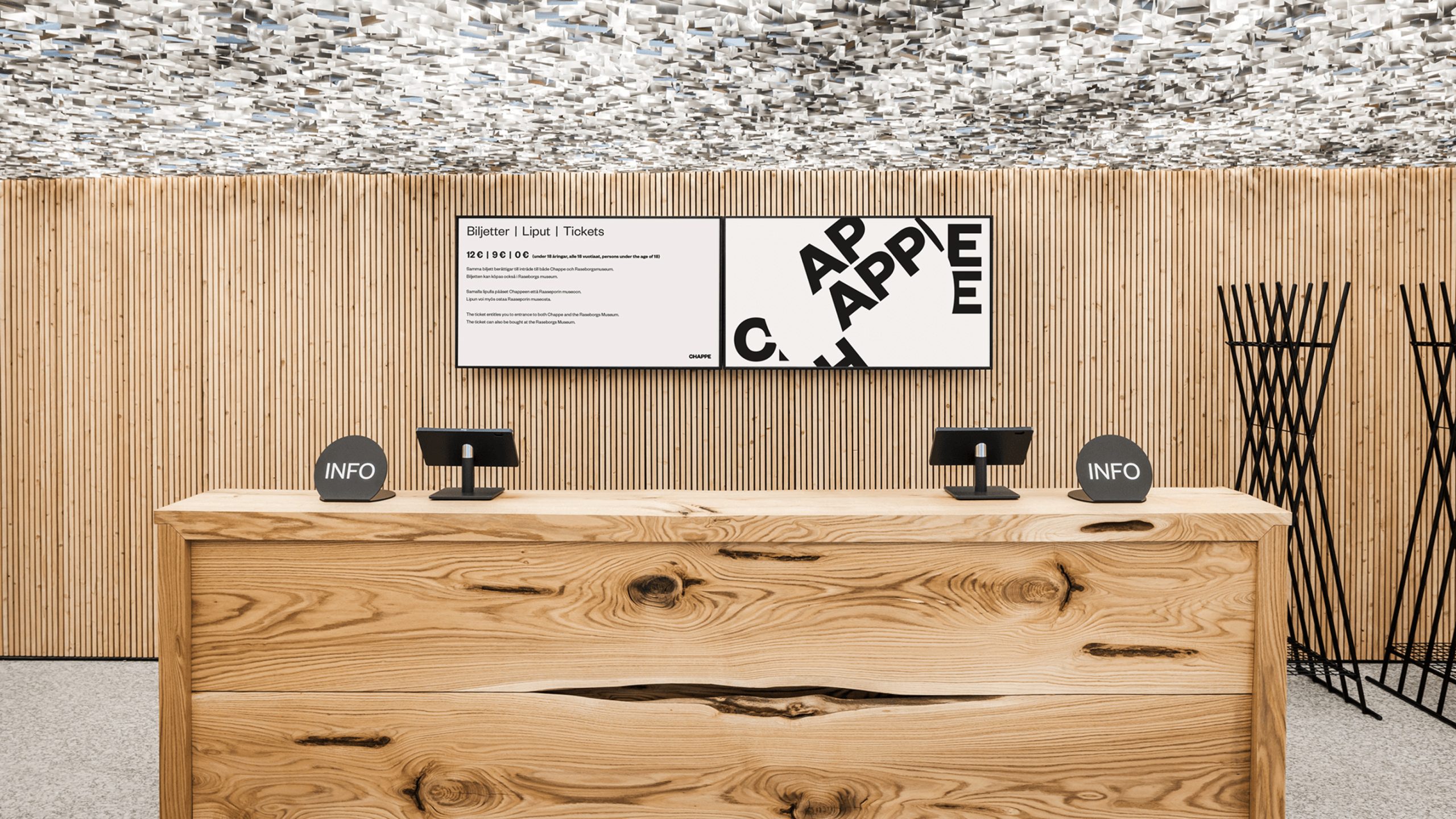 Chappe museum reception desk in natural wood with INFO signs, dual screens showing ticket prices, and a sculptural ceiling installation.