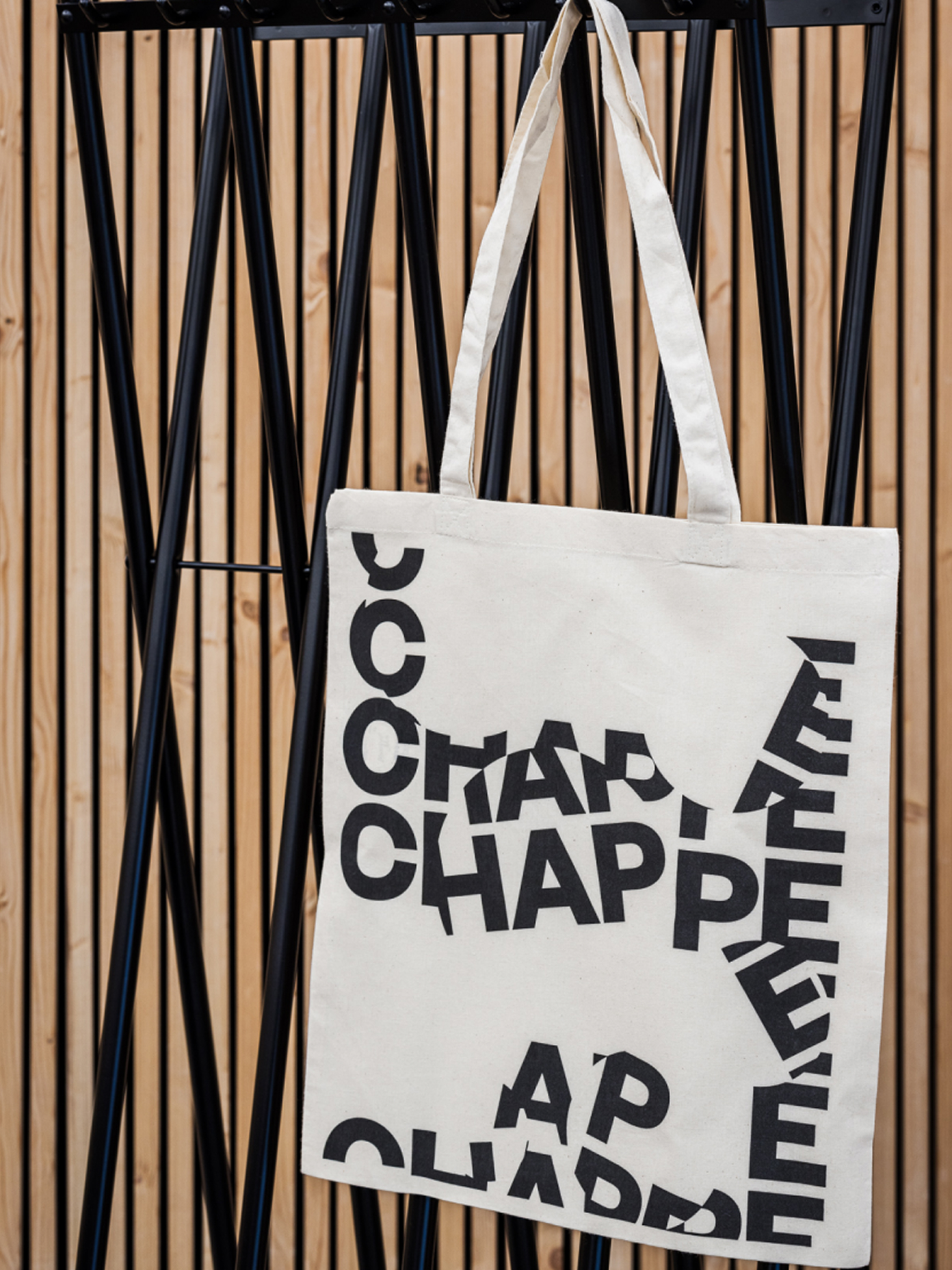 Cream canvas tote bag printed with repeating 'Chappée' text in bold black type, hung on a black metal rack.