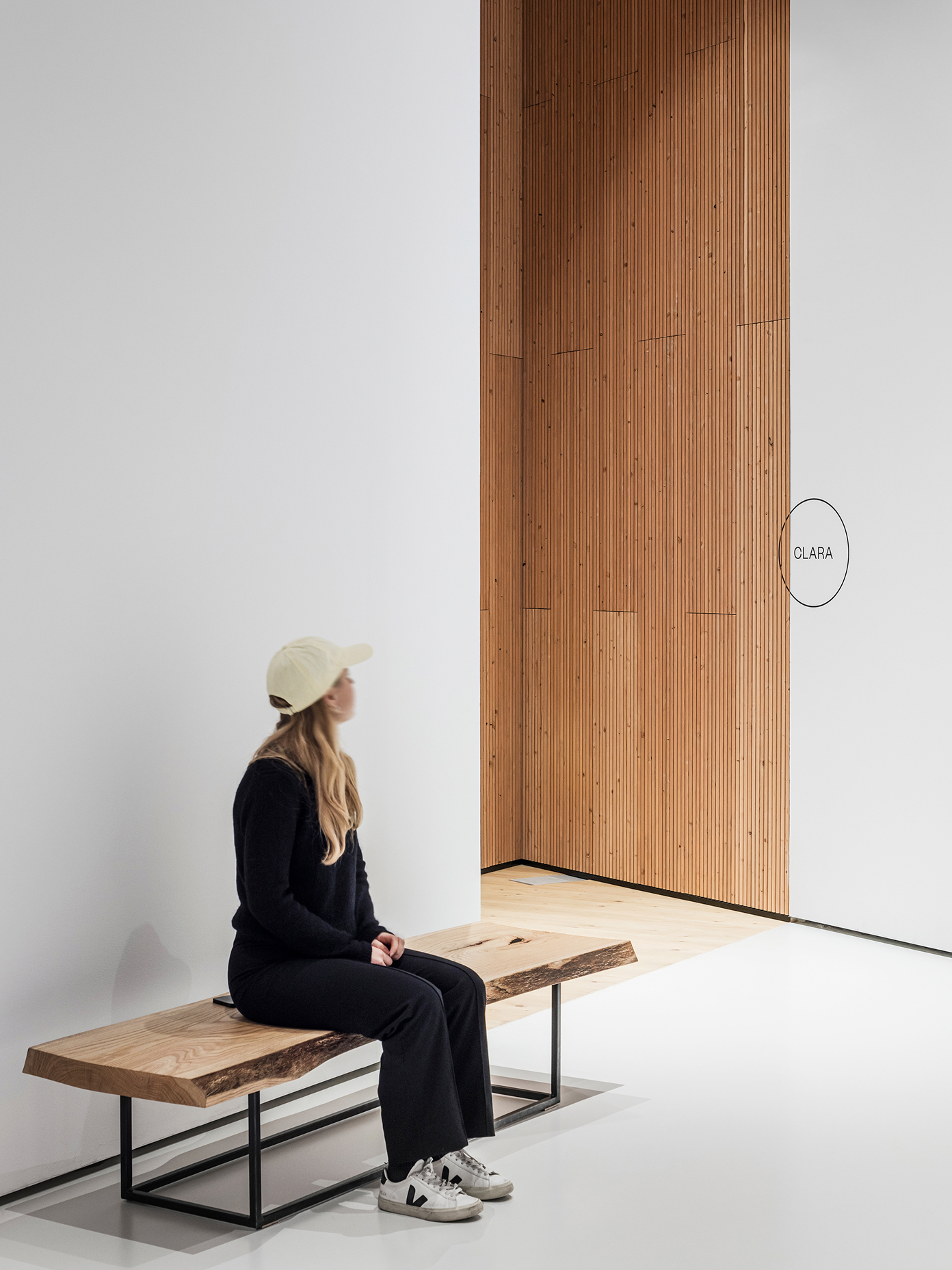 Woman in black outfit sits on a live-edge wood bench facing Chappe's slatted timber wall panel with circular brand signage.