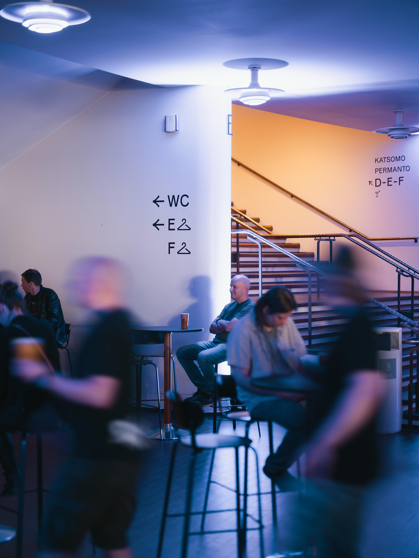 Kulttuuritalo foyer with motion-blurred visitors, directional signage, warm staircase lighting, and tiered seating area.