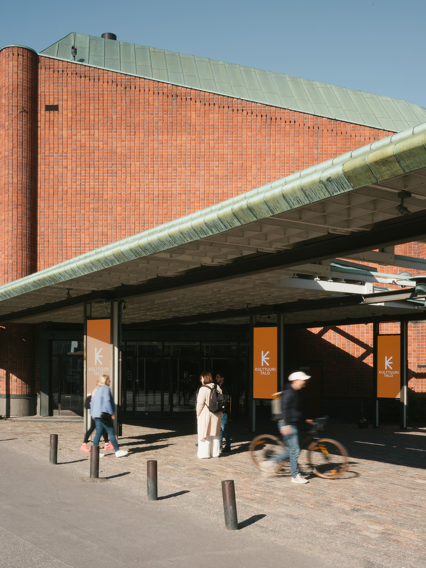 Kulttuuritalo entrance with red brick facade, green copper roof, and orange branded banners under a canopy in Helsinki.