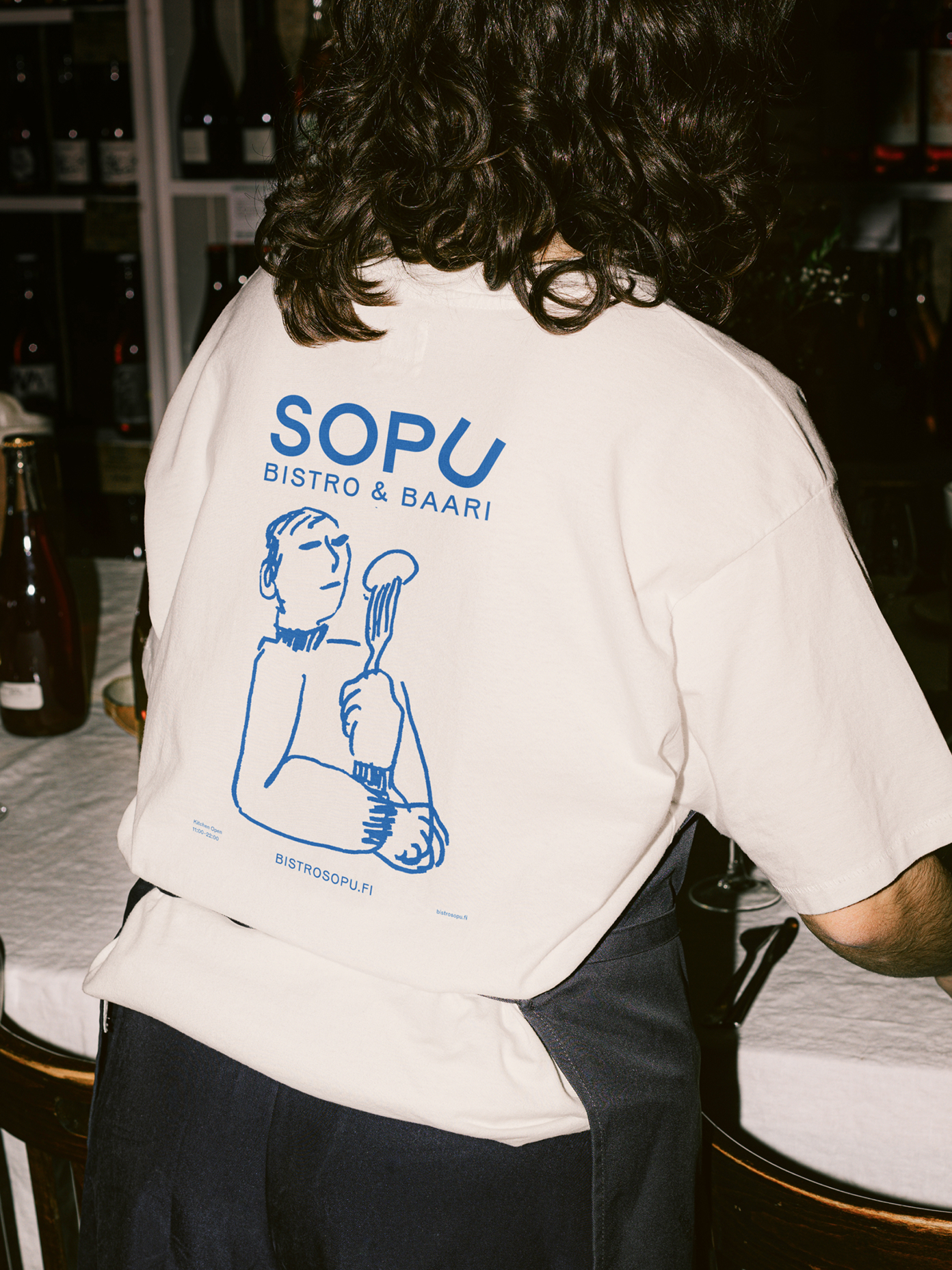 Back of a cream t-shirt with blue Sopu Bistro & Baari logo and illustrated figure holding cutlery.