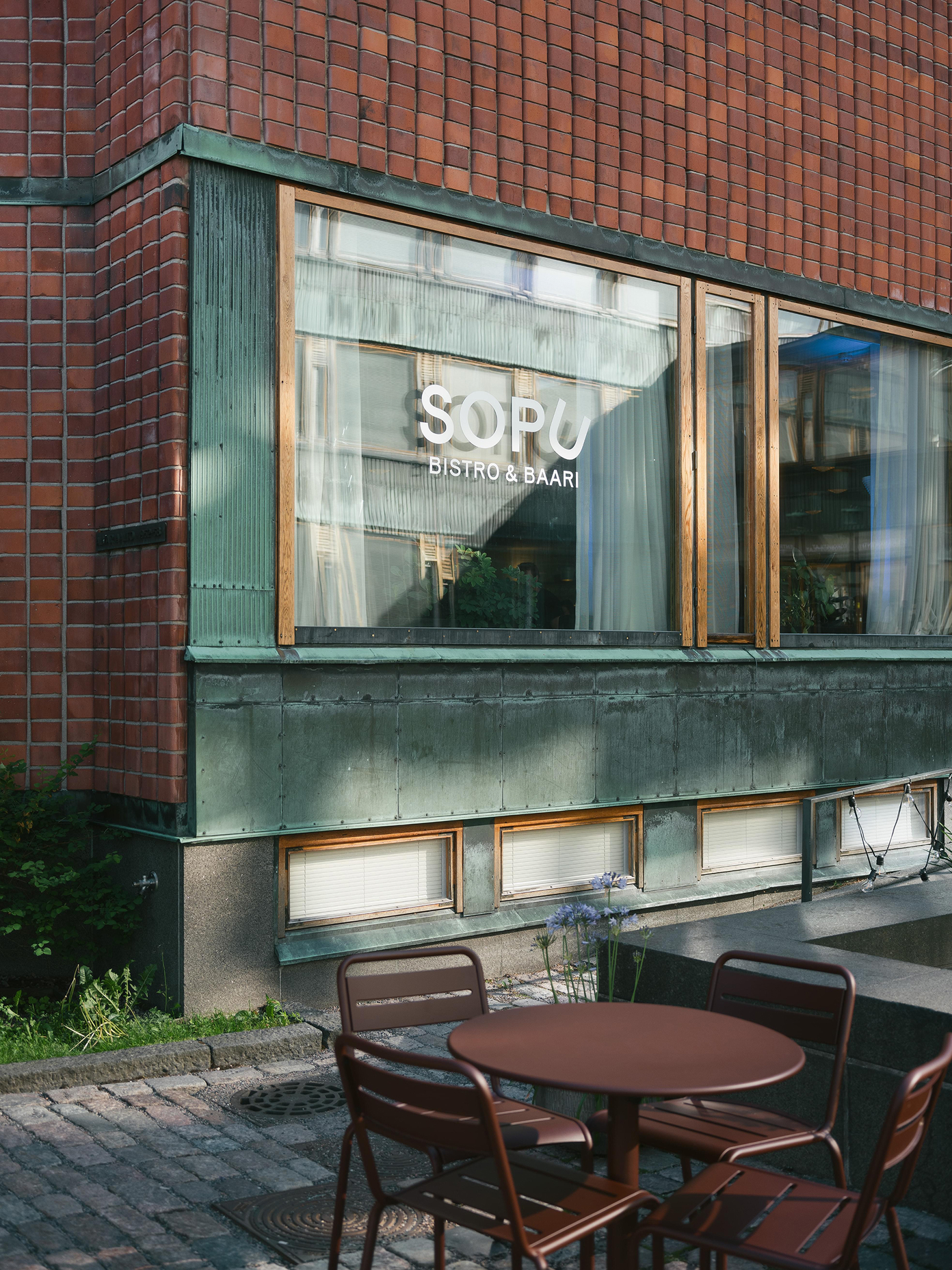 Sopu Bistro & Baari exterior with patinated copper cladding, large windows, and dark red outdoor seating on cobblestone.