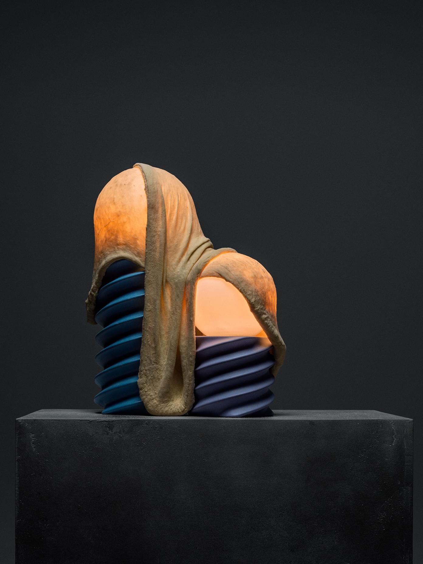 Glowing ceramic sculpture with draped skin-like texture over stacked blue discs, displayed on a dark concrete plinth.