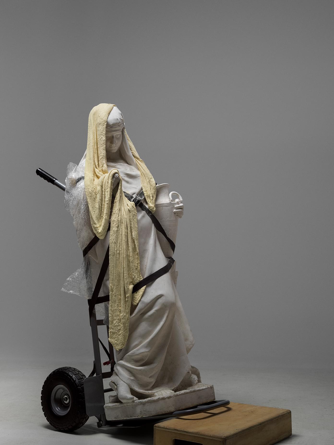 Classical white marble-style statue strapped to a hand truck with foam padding and bubble wrap for transport.