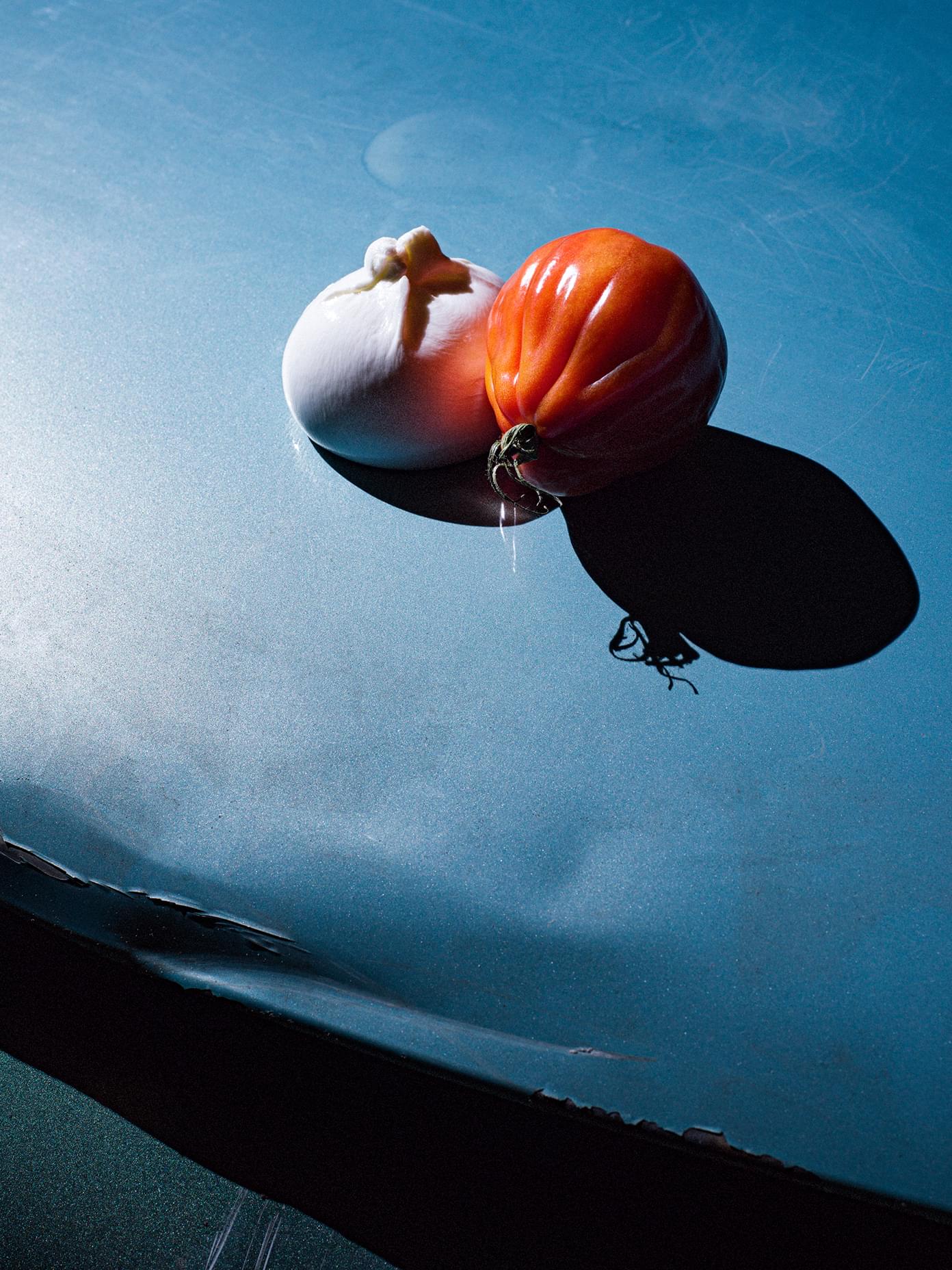 A burrata and heirloom tomato rest together on a blue surface, casting a dramatic shadow.