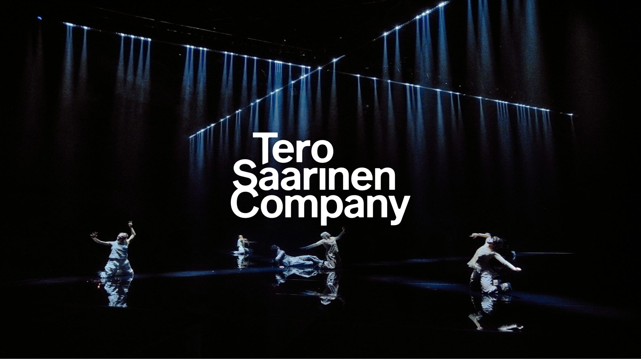 Tero Saarinen Company dancers perform on a dark reflective stage under dramatic blue beam lighting.