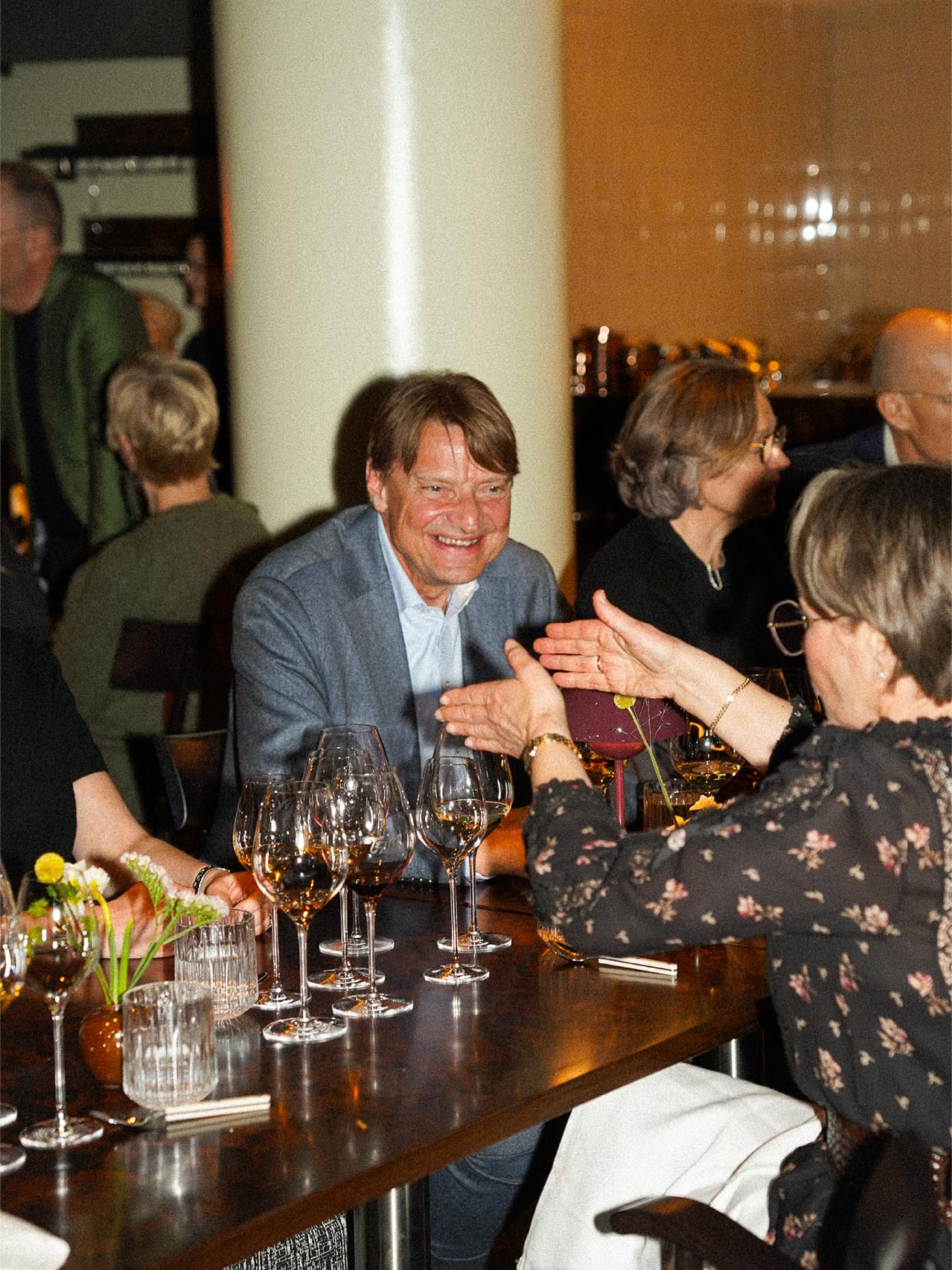 Guests laughing and clapping at a Teller dining event, wine glasses and small floral arrangements on a dark wooden table.