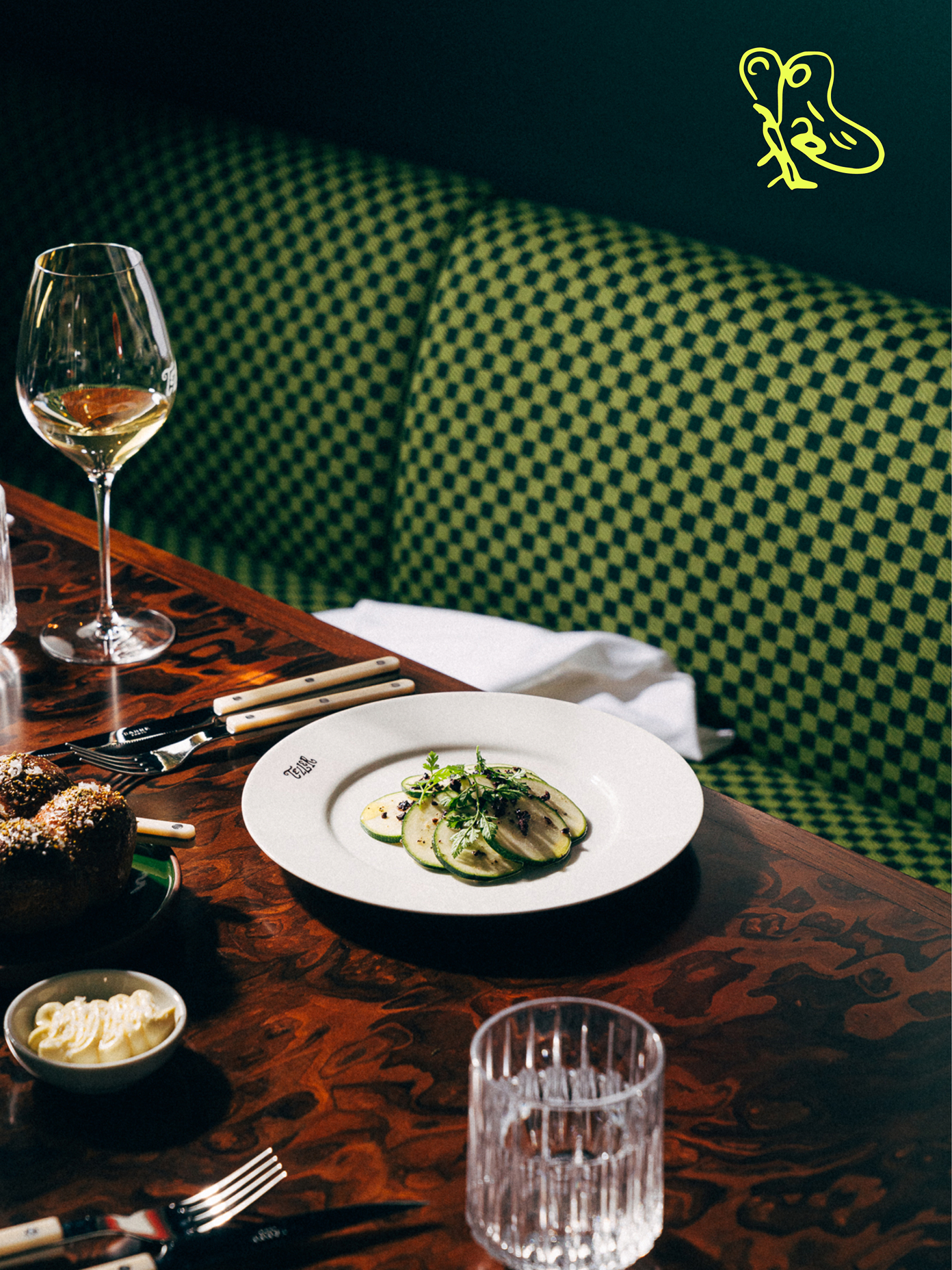 Teller restaurant table with zucchini carpaccio on branded white plate, white wine, dark bread, and green checkered booth seating.
