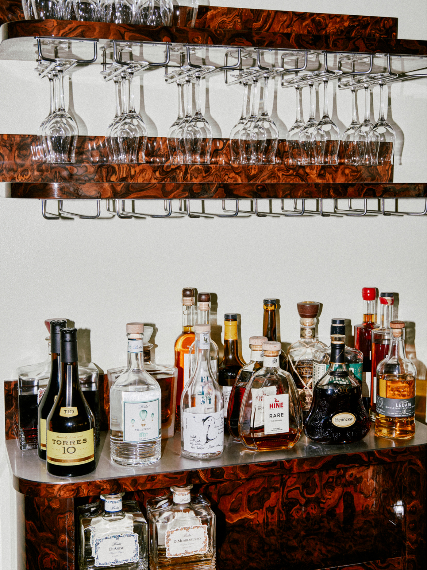 Teller bar cart with bottles beneath suspended wine glasses on burl wood shelving.