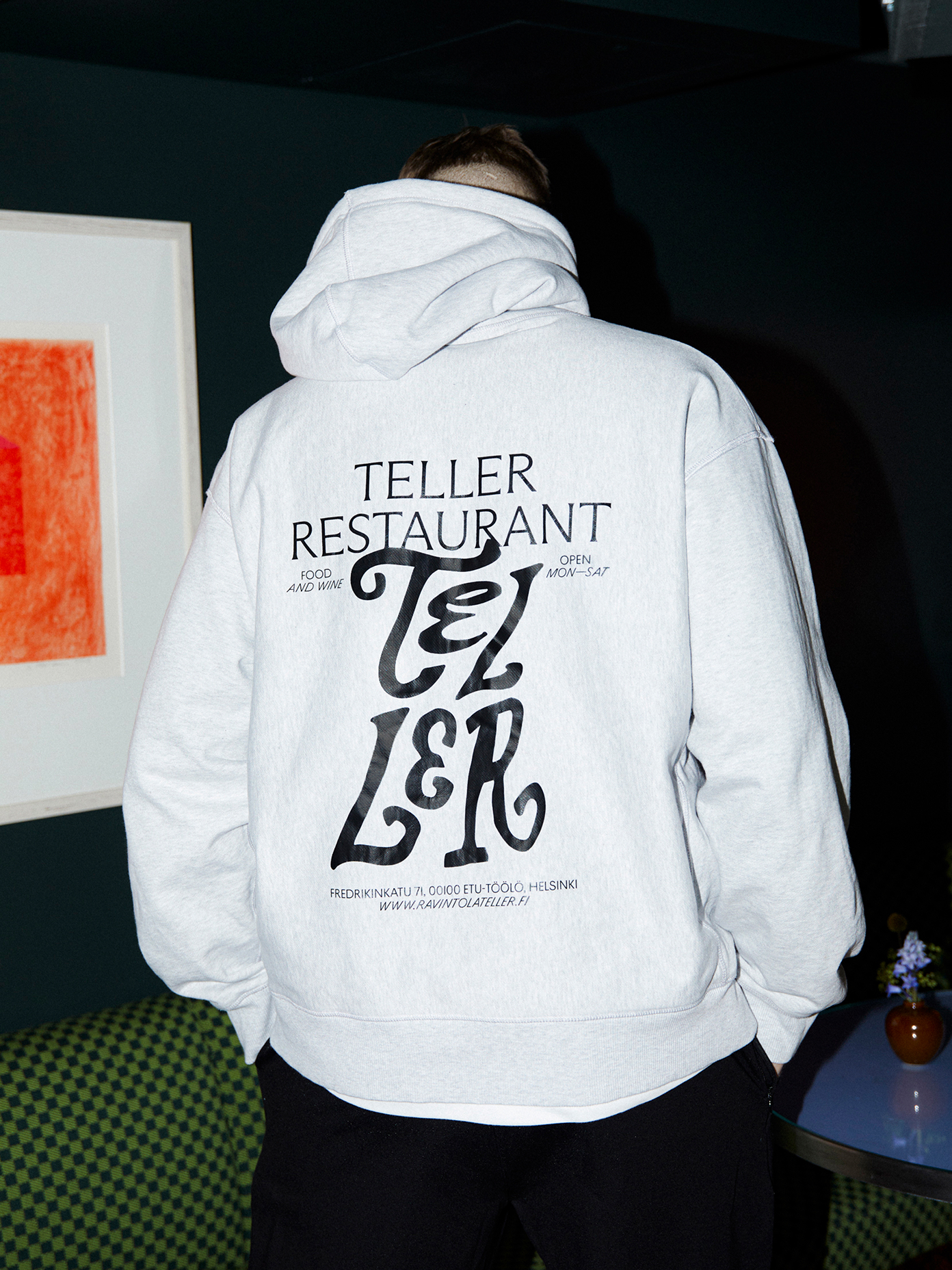 Person wearing a heather grey Teller Restaurant hoodie showing bold typographic back print with Helsinki address details.