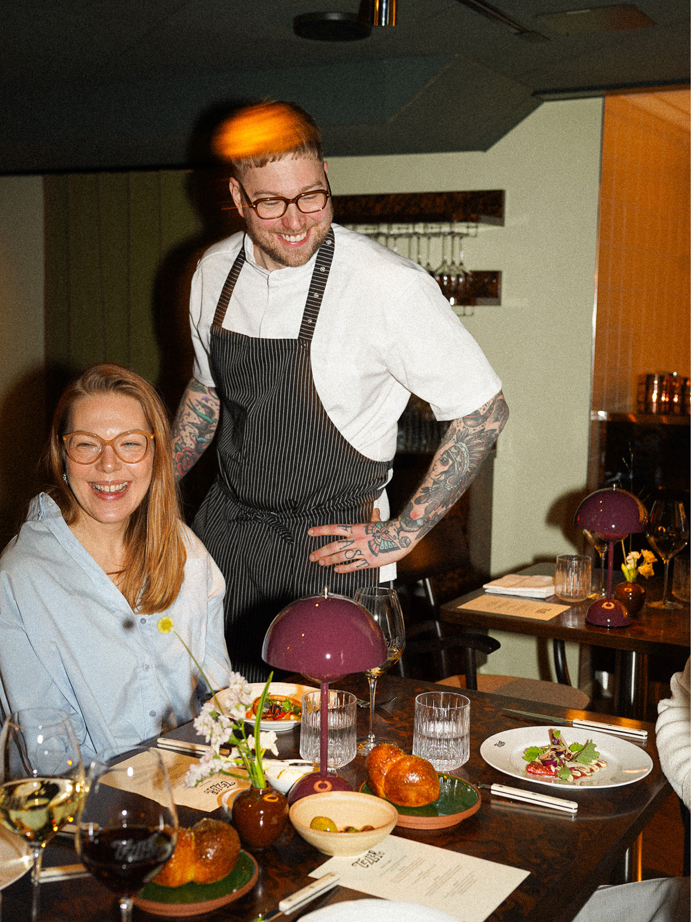 Chef in Teller apron smiles with a seated guest at a candlelit restaurant table set with elegant dishes.